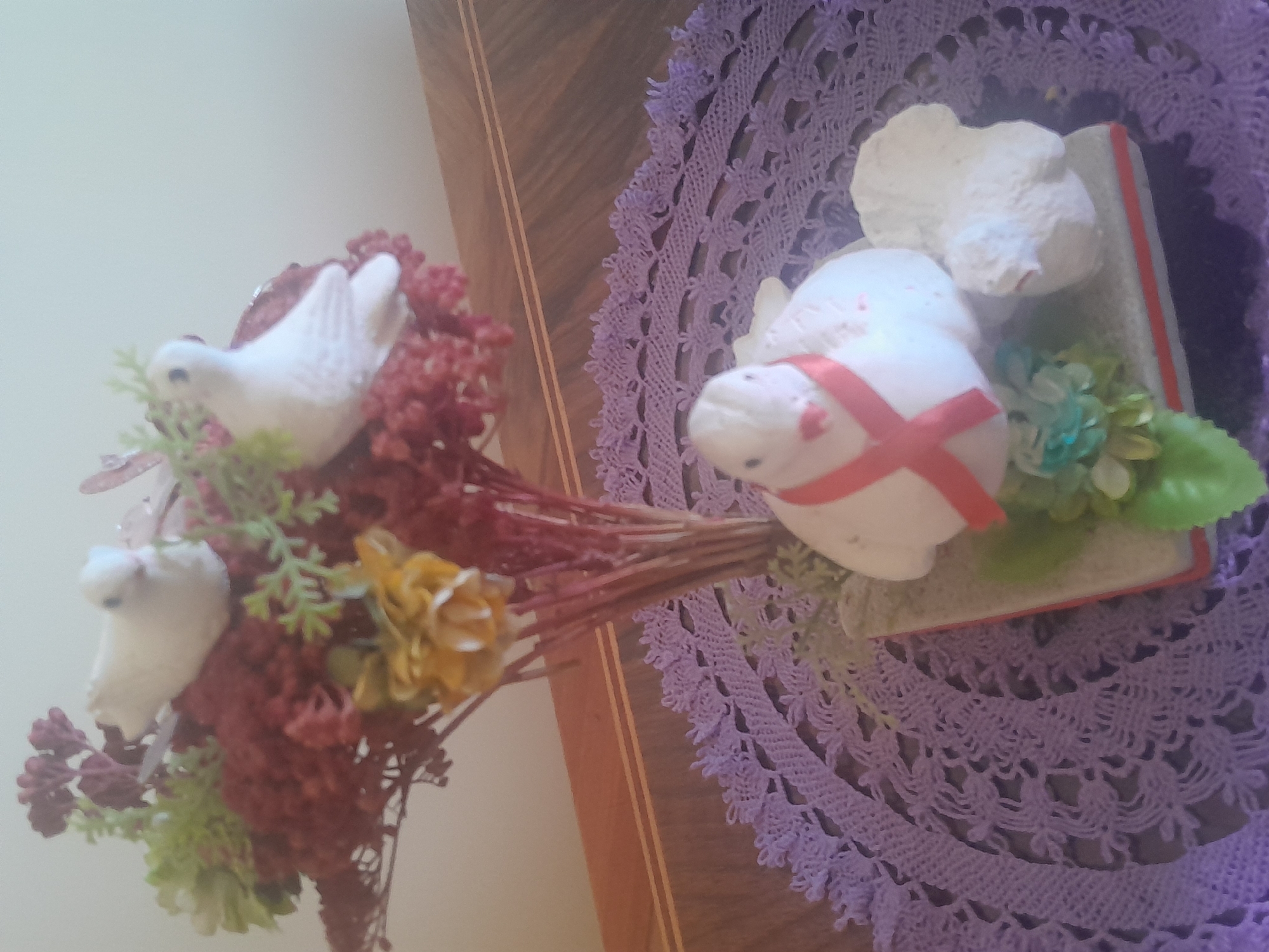 A still life arrangement is presented on a wooden table, partially obscured by a purple crocheted doily. The arrangement features a bouquet of artificial red and yellow flowers with green foliage, topped by two white ceramic doves. In front of the bouquet, a larger white ceramic dove sits on a rectangular base adorned with a red ribbon tied in a bow. Another smaller white ceramic figure, possibly a bird, is positioned to the right of the larger dove. The overall lighting is dim, casting a soft glow on the objects. The location is not specified in the image.