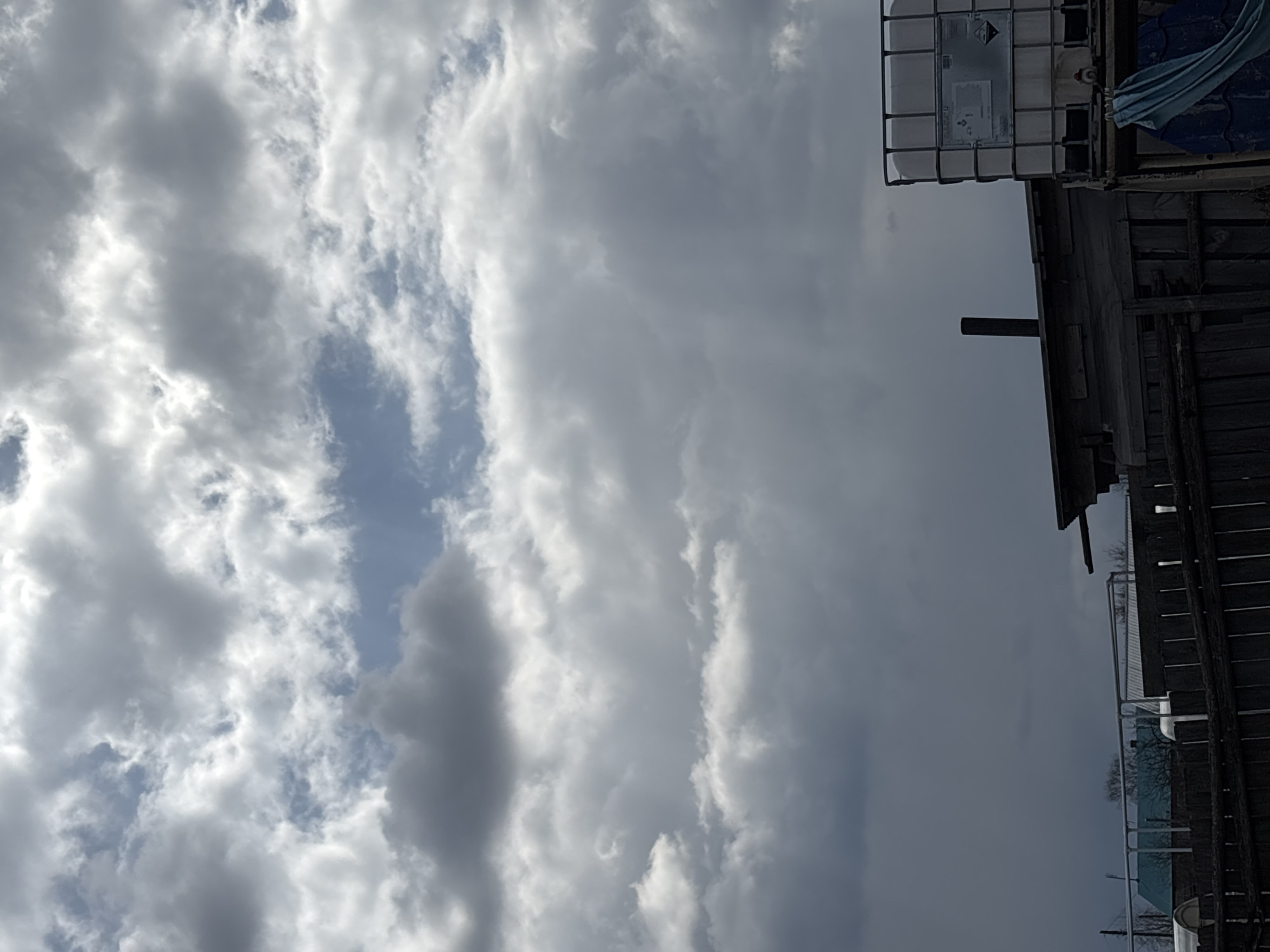 The image, taken in Фоки, Russia, depicts an outdoor scene dominated by a dynamic, partially cloudy sky. The sky features large, bright white and darker grey cumulus clouds, with patches of clear blue sky visible between them, indicating sunlight breaking through.
On the right side of the frame, various industrial or agricultural structures are visible. A prominent subject is a large, white intermediate bulk container (IBC tote) encased in a metal cage, positioned in the upper right. On its side, there is a visible label featuring a black GHS hazard pictogram, likely indicating a corrosive or environmentally hazardous substance, followed by some unreadable text. Below and to the right of the IBC tote, parts of rustic or industrial structures, possibly sheds or outbuildings made of wood and metal, are present. A dark, vertical pipe or vent protrudes from one of these structures. A piece of blue tarp or fabric is also draped over an object near the IBC tote. Further down on the right, more indistinct structural elements, possibly fences or additional building sections, are visible.
The scene is static, devoid of human presence or overt activity. The time of day appears to be daytime, given the visible sunlight and the clarity of the clouds. The setting suggests an industrial, agricultural, or rural storage area.