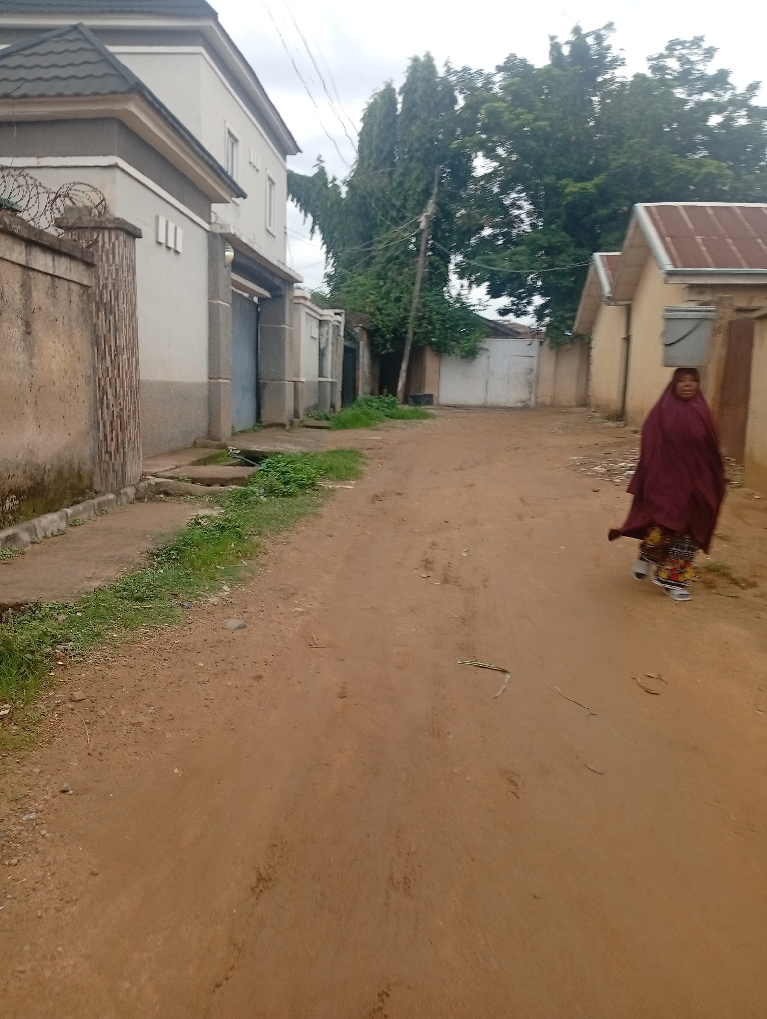 A woman, draped in a dark red hijab and skirt, walks along a dirt road in what appears to be a residential area of Rafin Par, Nigeria. The road is unpaved and dusty. To the left of the road, a light-colored, multi-story building with a dark gray roof is visible, bordered by a beige concrete wall topped with barbed wire. Adjacent to this building is a dark gray metal gate. Further down the road, to the right, a small, light yellow building with a corrugated metal roof stands. Behind the buildings, lush green trees dominate the background, with some power lines strung between poles. The sky is overcast.