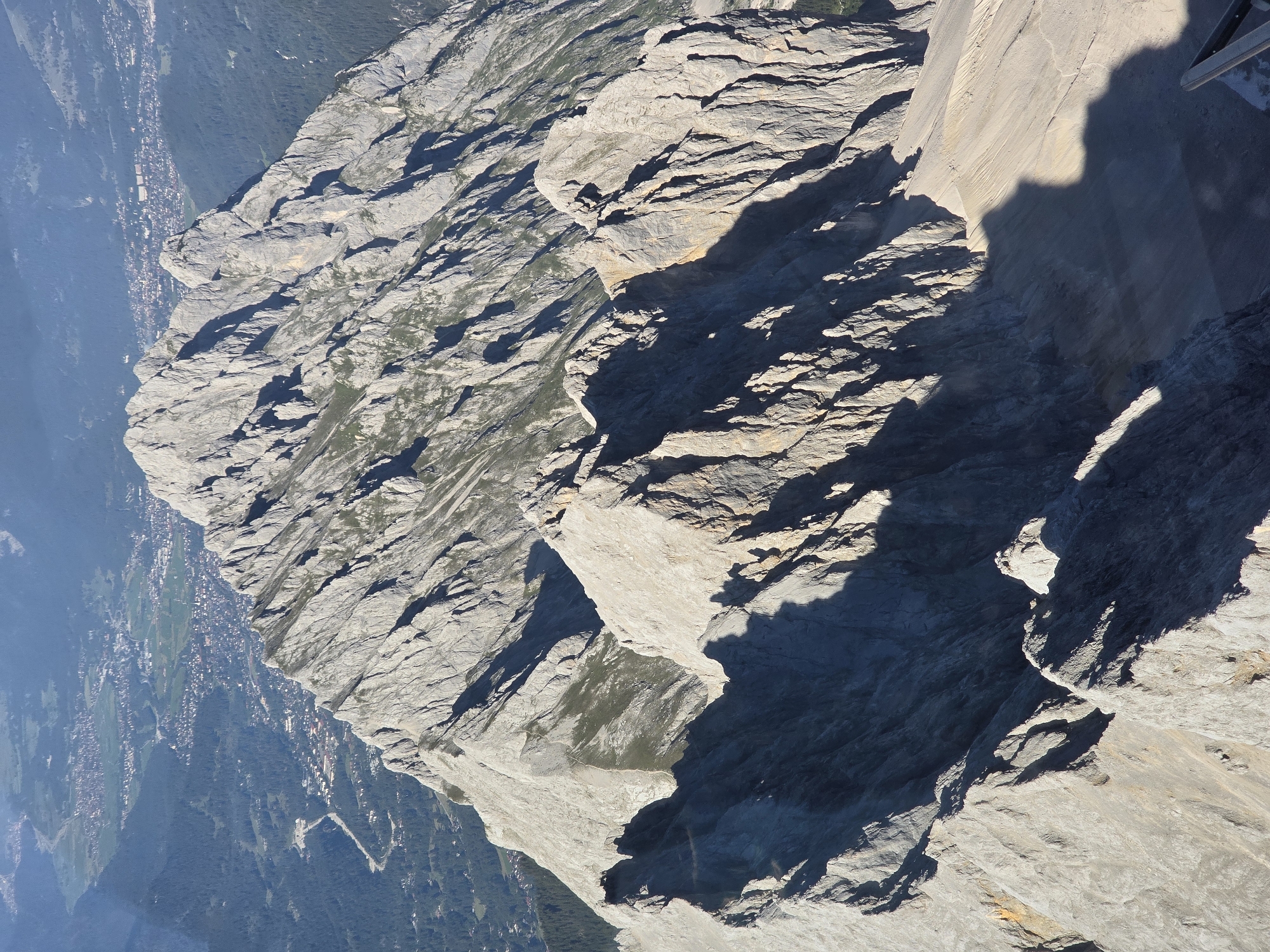 This is a high-angle aerial view of a rugged mountain range with a town nestled in the valley below, identified as Garmisch-Partenkirchen, Germany. The foreground is dominated by sharp, rocky peaks casting long shadows. The mountains are predominantly gray and appear to be composed of weathered rock, with visible striations and crevices. Patches of green vegetation are scattered across some of the gentler slopes.
In the mid-ground, the mountain ridges continue, leading the eye towards the valley. The town of Garmisch-Partenkirchen is visible in the distance, appearing as a collection of buildings with red roofs, interspersed with green spaces that suggest parks or fields. A winding road or river is discernible traversing the valley floor.
The background consists of further mountain ranges, appearing hazy and blue due to atmospheric perspective, with more distant settlements or infrastructure possibly visible on the slopes. The lighting suggests it is daytime, with the sun casting distinct shadows that accentuate the mountainous terrain. No people or specific objects that are not part of the natural landscape or the town are clearly identifiable in the image.