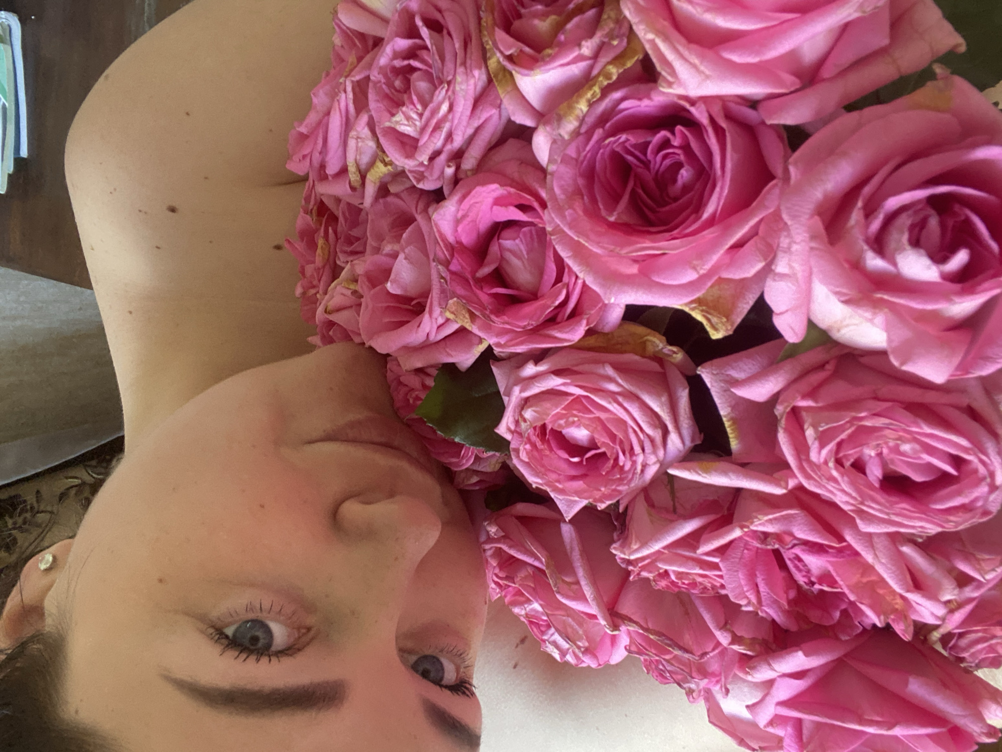 A close-up, slightly tilted photo shows a woman lying down, with her face visible in the lower left of the frame. A large bouquet of pink roses covers most of the right side of the image, partially obscuring her upper body. The woman has fair skin, light blue eyes, and dark eyebrows. An earring is visible in her left ear. The background is partially visible on the left side and appears to be a wooden surface with a patterned fabric beneath. The specific location in Sochi, Russia, cannot be determined from the provided image.