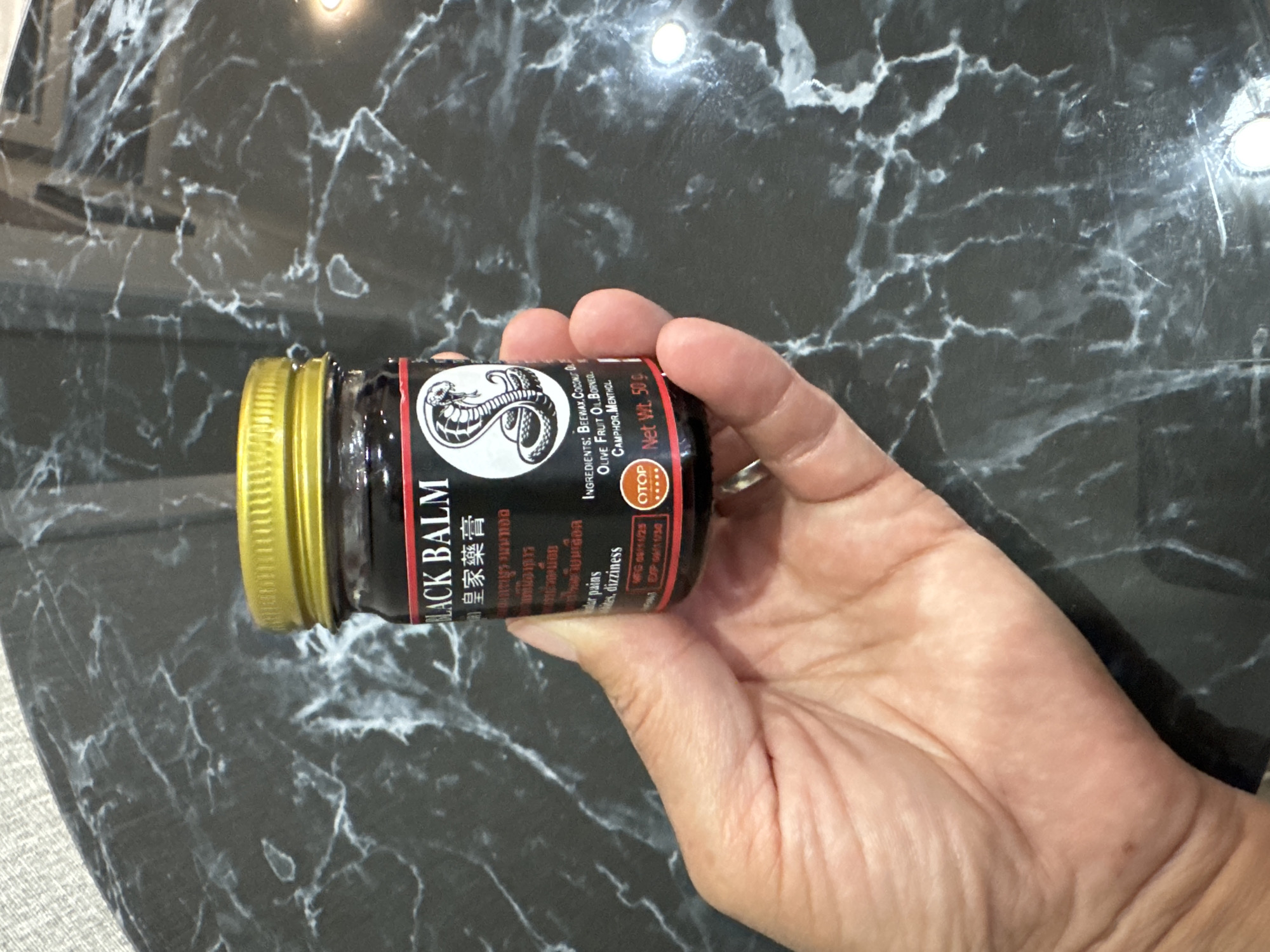 A hand holds a small, dark glass jar with a gold lid. The jar contains a dark balm and has a label with text in English and what appears to be Chinese characters. The label features an image of a cobra. Below the cobra image, the ingredients are listed as "BEEWAX, COCONUT OIL, OLIVE FRUIT OIL, BORNEOL, CAMPHOR, MENTHOL." The net weight is indicated as "50g". The jar is placed on a dark, marbled surface. The scene is indoors, with the marbled surface appearing to be a tabletop. The specific location in the city of Солонешное, Russia is not discernible from the image.