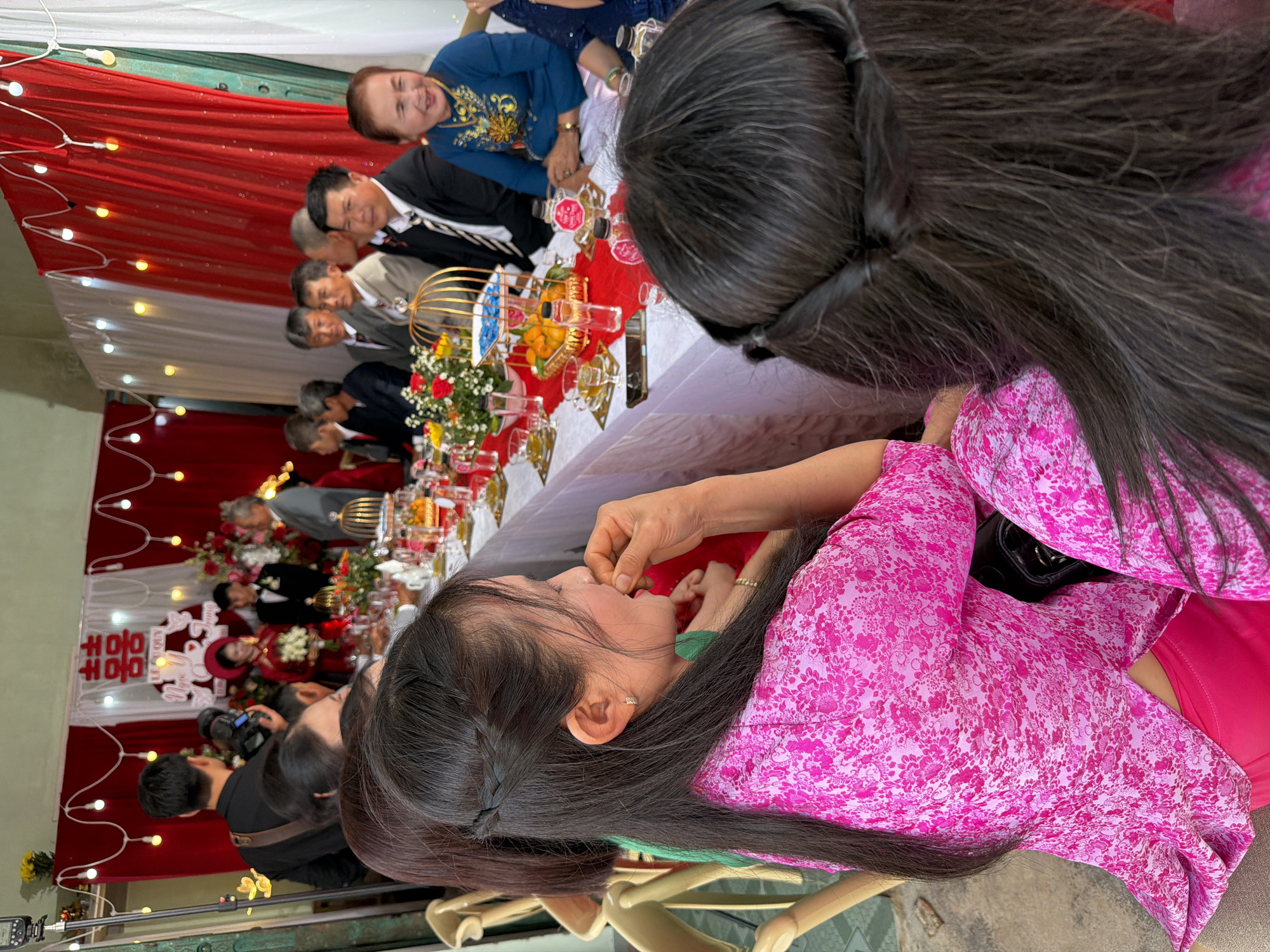 This media file depicts a vibrant wedding reception or engagement ceremony taking place in Tay Son Commune, Vietnam. The setting features decorative red and white fabric drapes, creating a festive atmosphere, illuminated by strands of small, warm-toned fairy lights.
In the immediate foreground, a woman with long, dark hair, styled with a side braid, is partially visible. She is wearing a striking pink floral-patterned long-sleeved top over a green inner garment and is captured in the act of bringing something to her mouth, possibly food. To her right, the back of another person's dark-haired head is seen.
Further into the scene, a long table is centrally arranged with a white tablecloth and a red runner. It is adorned with various celebratory items including small floral arrangements featuring red roses and white flowers, golden birdcage-like decorations, numerous small glass cups, and bowls of fruits such as oranges. Several guests are seated around this table; among them, a woman in a blue traditional Vietnamese áo dài top is smiling broadly. Multiple men in suits and formal shirts are also seated, observing the event or interacting.
In the background, a focal point is a red and white backdrop, prominently displaying the red Chinese character "囍" (double happiness), which is a common symbol for weddings in Vietnamese and Chinese cultures. Below this, Vietnamese text is partially visible, likely indicating the type of ceremony, such as "LỄ ĂN HỎI" (engagement ceremony) or "LỄ VU QUY" (wedding ceremony). A couple, presumably the bride and groom, dressed in traditional attire, are positioned near this backdrop. A photographer is also present, capturing the event with a camera mounted on a tripod, suggesting the formal documentation of this joyous occasion. The overall mood conveyed by the smiling faces and festive decor is one of celebration and happiness.