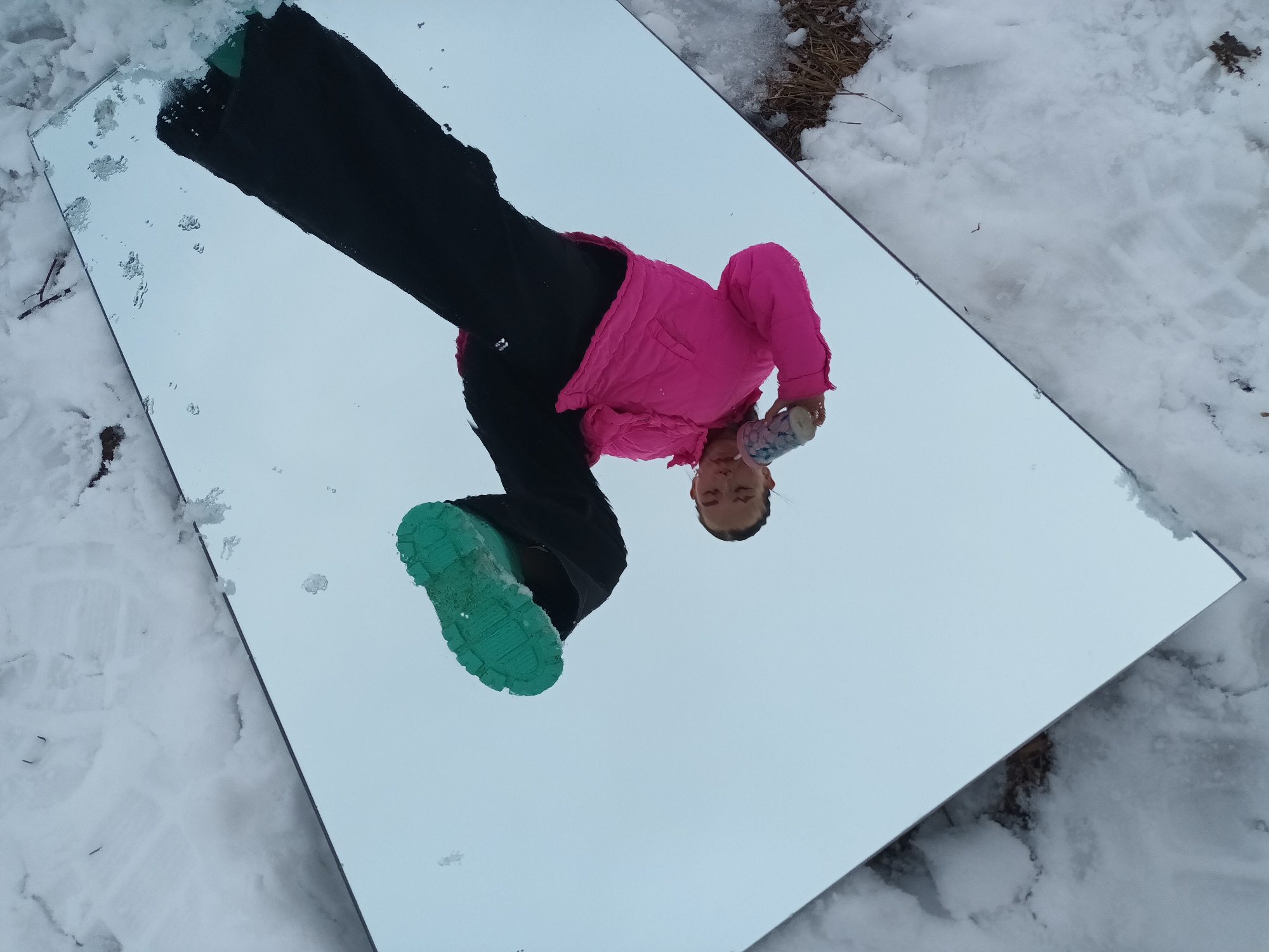 A girl is standing on a mirror placed on the ground in a snowy outdoor environment. The girl is wearing a pink jacket, black pants, and teal-soled shoes. She is holding a container in her right hand. The mirror reflects the sky, giving the illusion that the girl is floating. The ground around the mirror is covered in snow, with some patches of brown vegetation visible. The image was taken in Ольховка, Russia.