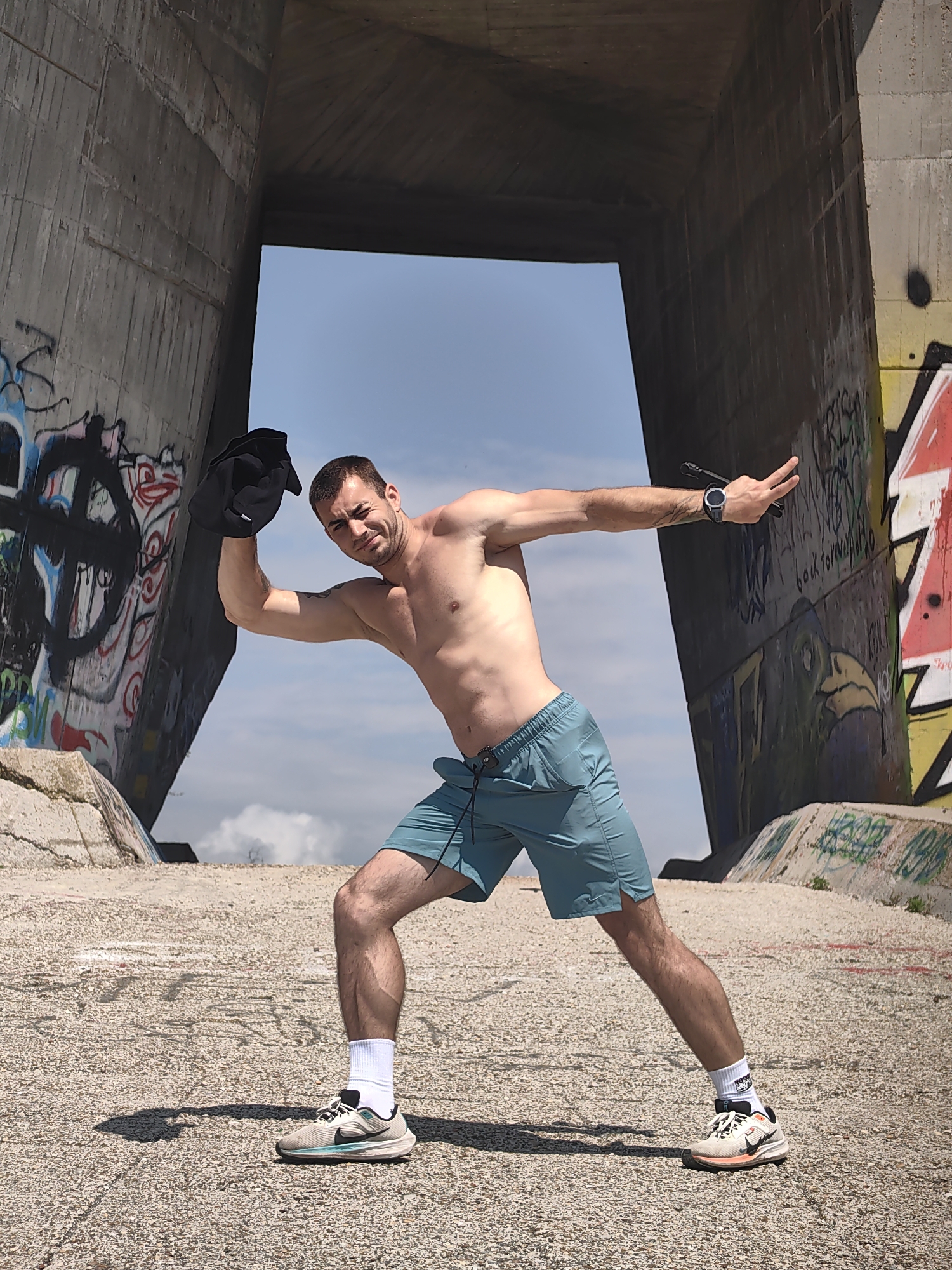 An adult male, approximately 20-30 years old, with short brown hair and light stubble, stands shirtless in a dynamic, low stance within a concrete architectural structure in Varna, Bulgaria. He is wearing teal athletic shorts, white socks, and athletic shoes. A black sports watch is visible on his left wrist, and a tattoo adorns his left forearm. His right arm is bent upwards, holding a black garment or cap, while his left arm is extended straight outwards, palm open. He looks directly at the camera with a focused expression. The setting is an outdoor, light-colored pebbled concrete surface framed by two massive, angled concrete walls that form an overhead archway or underpass. Both concrete walls are heavily covered in graffiti; the left wall features large black, white, and red tags, while the right wall displays multi-colored graffiti, including a stylized bird figure and text reading "back forever." Bright blue sky with scattered white clouds is visible through the opening of the concrete structure, indicating clear daylight. Shadows are cast by the man on the ground and by unseen objects on the right wall.