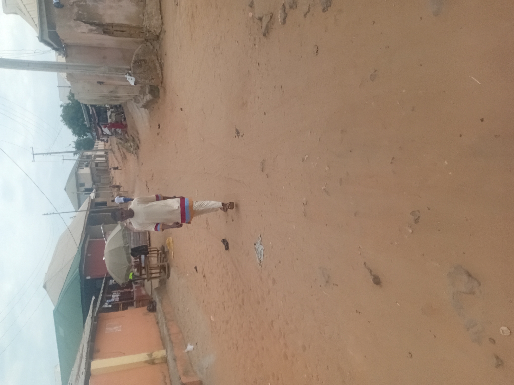 A person is walking on a dirt road in an urban area. The environment includes buildings of varying construction and age, an umbrella-covered structure possibly indicating a street vendor's setup, and visible power lines overhead. The color of the dirt suggests a dry climate or a lack of recent rainfall.