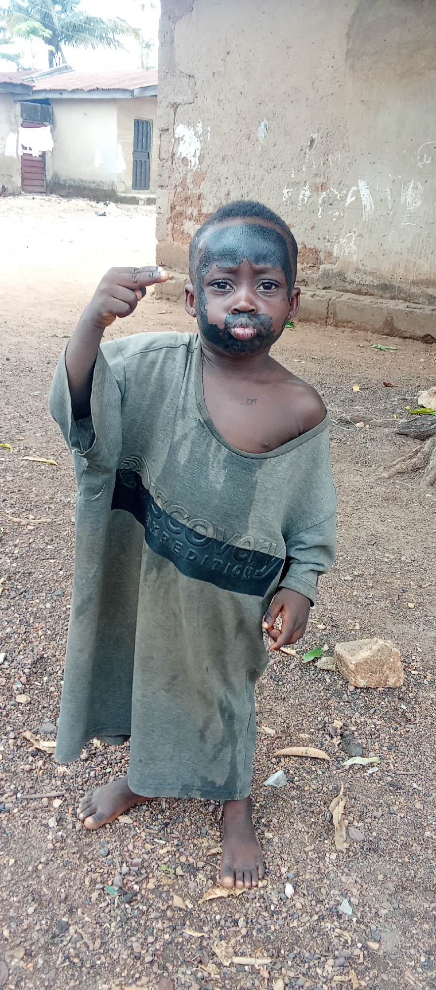 A young child stands barefoot on a rocky, dirt-strewn ground in Akwanga, Nigeria. The child's face is covered in a dark substance around the forehead and mouth. The child is wearing an oversized, stained, long-sleeved grey shirt. The child is making a gesture with the right hand raised, the index and middle fingers extended. Behind the child, there's a simple building with a corrugated iron roof, a wall, and a closed door. A second building is to the left. The ground has scattered stones and dry leaves. The lighting is daylight.
