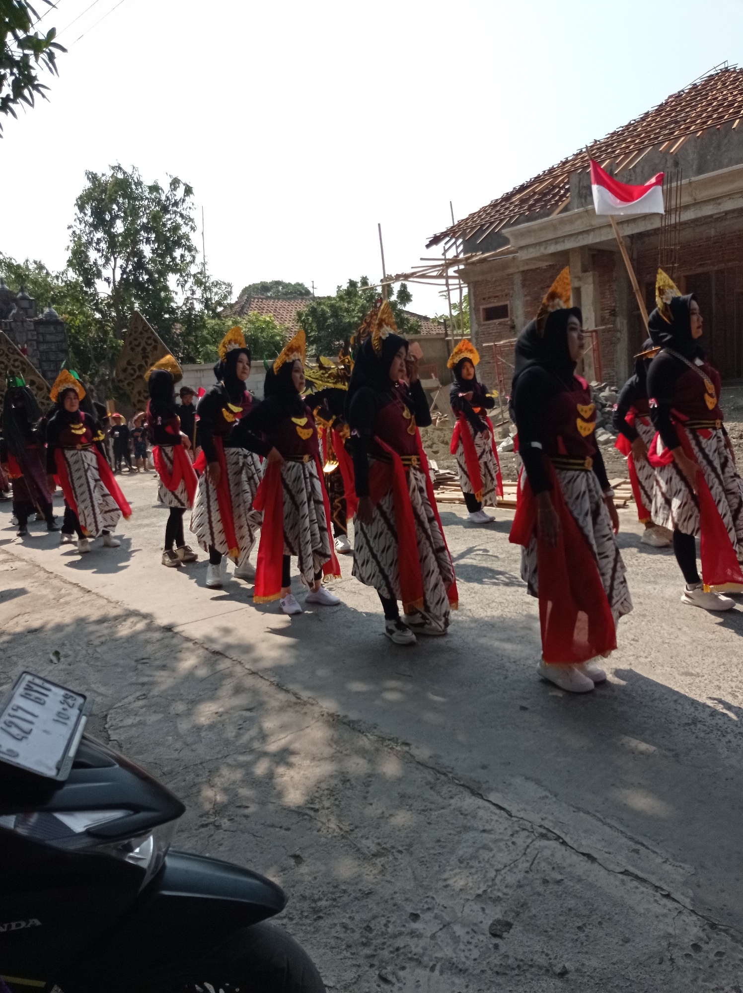 In Maribaya, Indonesia, a group of women, likely performers, are walking down a paved street under a clear, bright sky. They are dressed in traditional attire consisting of black tops, red sashes, and patterned skirts with red accents. Many of the women are wearing ornate headpieces, and some have gold necklaces. The women are walking in a line, facing right, and appear to be participating in a parade or cultural event. In the background, a partially constructed building with a tiled roof is visible, and an Indonesian flag is flying from a pole in front of it. There are trees and other vegetation in the background as well. In the foreground, the front of a dark-colored motorcycle is visible, with a license plate that reads "G 4217 BYF 10-29." The street is lined with shadows cast by the surrounding trees and possibly the people in the procession.