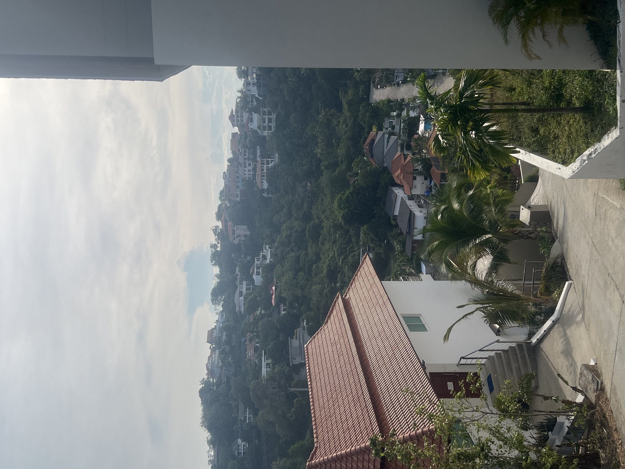 The image displays a view from an elevated position overlooking a hillside residential area in Ko Samui District, Thailand. In the foreground, a white building with a red tiled roof is partially visible. Concrete pathways and stairs lead away from this building and descend into a lush, green environment. Scattered across the hillside are numerous houses, many with white exteriors and red or grey tiled roofs, nestled amongst dense foliage and trees. A road winds through the lower part of the scene, with more houses visible on either side. Palm trees are prominent in the landscape. The sky above is partially clouded, with some sunlight breaking through. There are no people visible in the image.