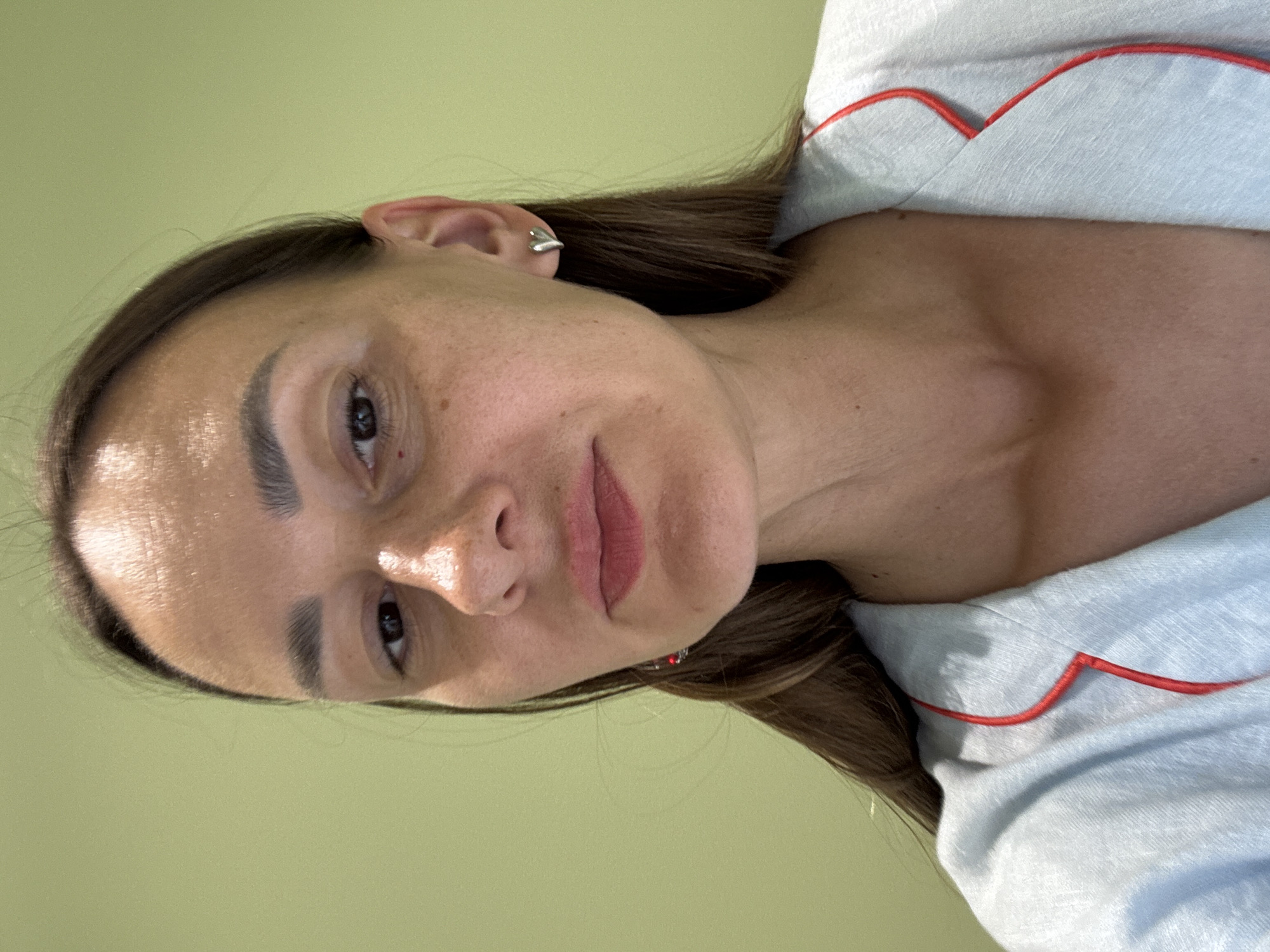 A medium shot captures a woman's face and upper body against a pale green background. She is looking directly at the camera with a neutral expression. The woman has long, dark brown hair that is straight and parted to her left, framing her face. Her eyebrows are dark and well-defined. She has a fair complexion with some subtle freckles visible on her cheeks and forehead. Her eyes are dark brown, and her nose is straight. Her lips are slightly parted, revealing a hint of her teeth, and appear to be a light pink color. She is wearing a light blue garment with red piping around the collar. A small silver heart-shaped earring is visible on her right ear. There is a hint of red on her left earlobe, suggesting another earring. The lighting suggests an indoor setting, possibly illuminated by overhead lights, as evidenced by the slight shine on her forehead. The overall impression is that of a casual selfie or a portrait taken in a relaxed environment. There is no indication of the city of Sochi, Russia, in the image.