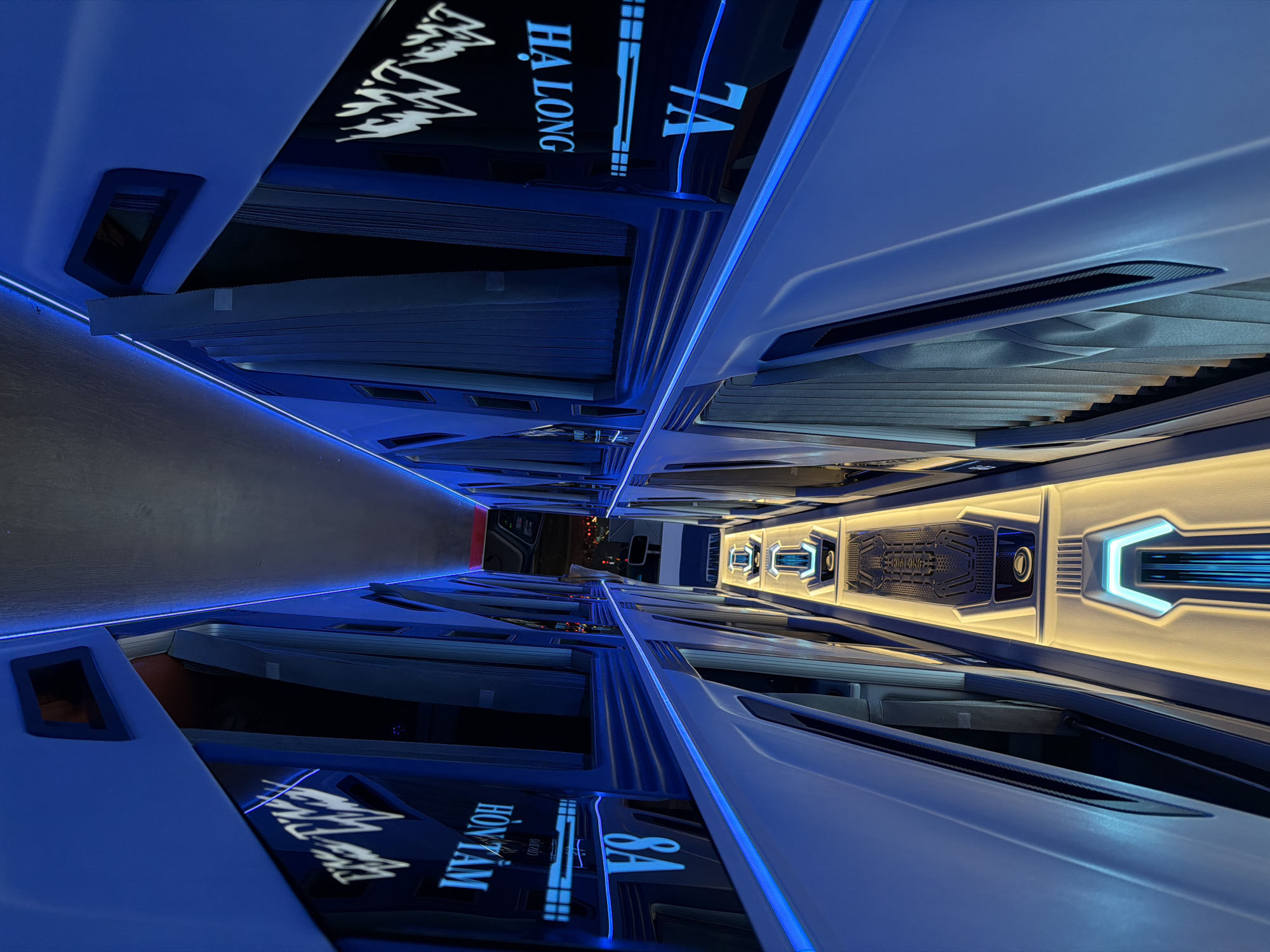 The image displays the interior of a modern sleeper bus, characterized by a sleek, futuristic design. The perspective looks down a central aisle, flanked on both sides by individual, capsule-like berths designed for rest during travel.
The primary lighting source is cool blue LED strip lighting that traces the edges of the berths and runs along the floor, contributing to a serene and high-tech ambiance. In contrast, the central ceiling features panels with warmer, yellow-white backlighting and decorative, angular fixtures that also incorporate blue light accents.
Each berth includes privacy curtains, visible in a pleated design. Above some of these berths are dark, reflective panels that display illuminated text. On one side, "HÀ LONG" (referring to Ha Long Bay) is clearly visible alongside "7A," likely a berth number. On the opposing side, "HÒN TẰM" (Hon Tam Island) and "8A" are shown. A central overhead panel also features the text "KIM LONG," which is a common name for bus companies in Vietnam.
No people are present in the scene. The artificial lighting and the dim, indistinct view through the windows at the far end of the aisle suggest the image was taken at night or during low-light conditions, possibly during a long-distance journey. The design emphasizes passenger comfort and privacy, indicative of a luxury travel experience in Vietnam.