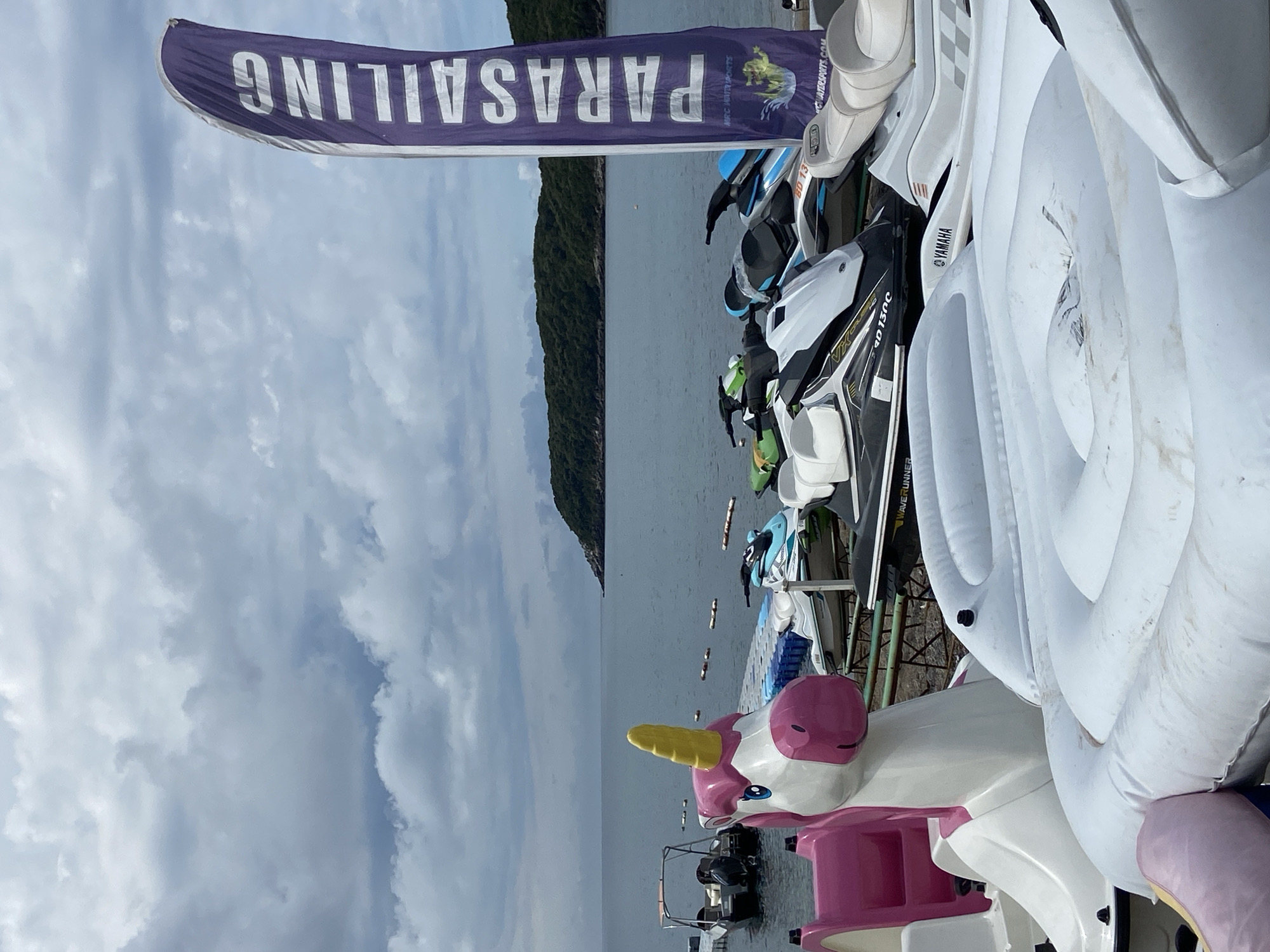 The image shows a waterfront scene in Prijevor, Montenegro. In the foreground are various watercraft: a unicorn-shaped paddle boat, several Yamaha jet skis, and inflatable towables. A purple banner with the word "PARASAILING" is visible in the upper left. In the background, the sea stretches out with a wooded coastline visible across the water. Several small boats are scattered on the water. The sky is overcast with cloudy conditions.