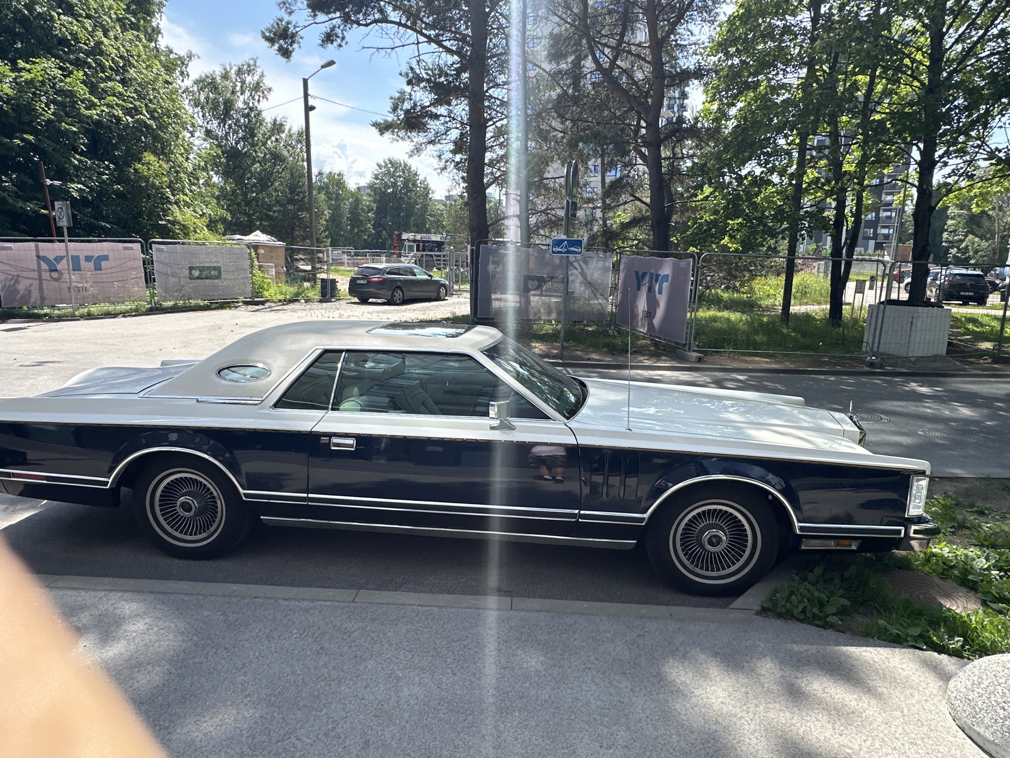 A dark blue and silver two-door Lincoln Continental Mark V is parked on a paved surface next to a sidewalk, with a construction fence in the background. The car's exterior is clean and reflective, showing the chrome trim and the distinctive opera windows. Beyond the fence, there are trees, a street lamp, and buildings, suggesting an urban environment in Tallinn, Estonia. A dark gray car is visible on the other side of the construction site. The sky is bright blue with scattered clouds. The scene appears to be illuminated by bright daylight, with some lens flare present in the image.