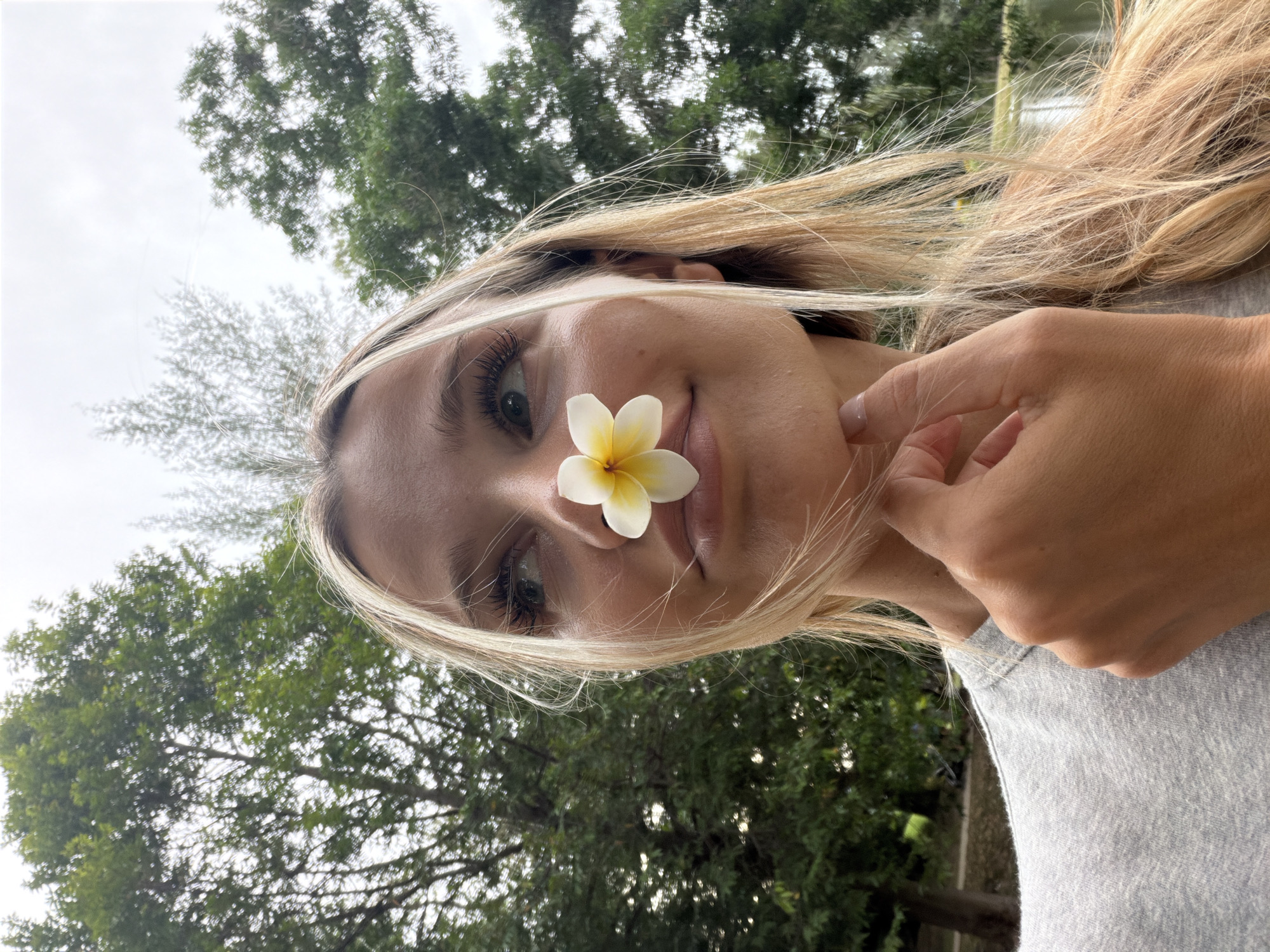 A young woman with blonde hair is photographed from the chest up, with a white and yellow flower placed on her nose. She is outdoors, with lush green trees and a body of water visible in the background. The sky is overcast. Her right hand is raised to her chin, and her head is tilted to the left as she looks upward. She is wearing a grey t-shirt.