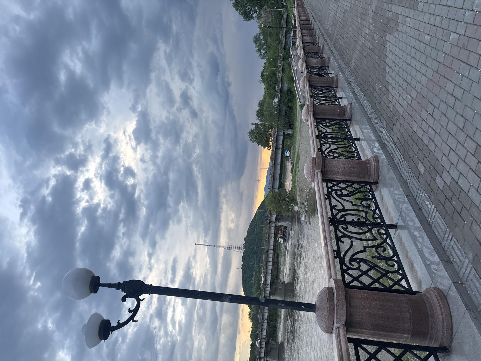 The image depicts a waterfront promenade in an unspecified location. A stone-paved walkway runs along the right side of the frame, bordered by a decorative metal and stone railing. Beyond the railing, a river flows, with a bridge spanning across it in the middle distance. Hills covered in greenery and a tall tower are visible on the far bank. The sky is overcast with dramatic clouds, suggesting either early morning or late evening light. A street lamp with two globes stands prominently in the foreground on the left. There are no people or animals visible in the scene. The overall atmosphere is serene and somewhat moody due to the cloudy sky.