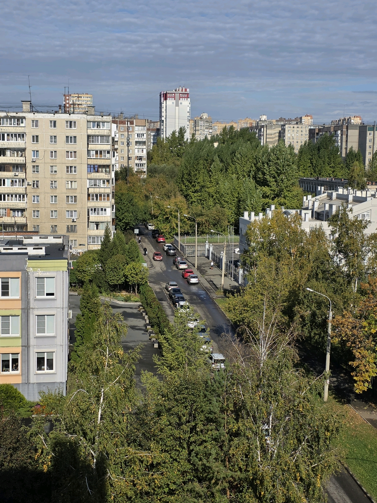 The image shows a street scene in a residential area of Chelyabinsk, Russia, on a partly cloudy day. In the foreground, a tall apartment building with light-colored facades and numerous windows is visible on the left. Adjacent to it is a lower apartment building with a gray and orange facade. A paved street runs through the middle of the image, lined with trees and parked cars. Several cars, including red, white, and dark-colored vehicles, are driving or parked along the street. People are walking on the sidewalk. In the background, more apartment buildings and trees are visible, extending into the distance under a sky with scattered clouds. The overall scene depicts a typical urban environment with residential buildings, a street, and greenery.