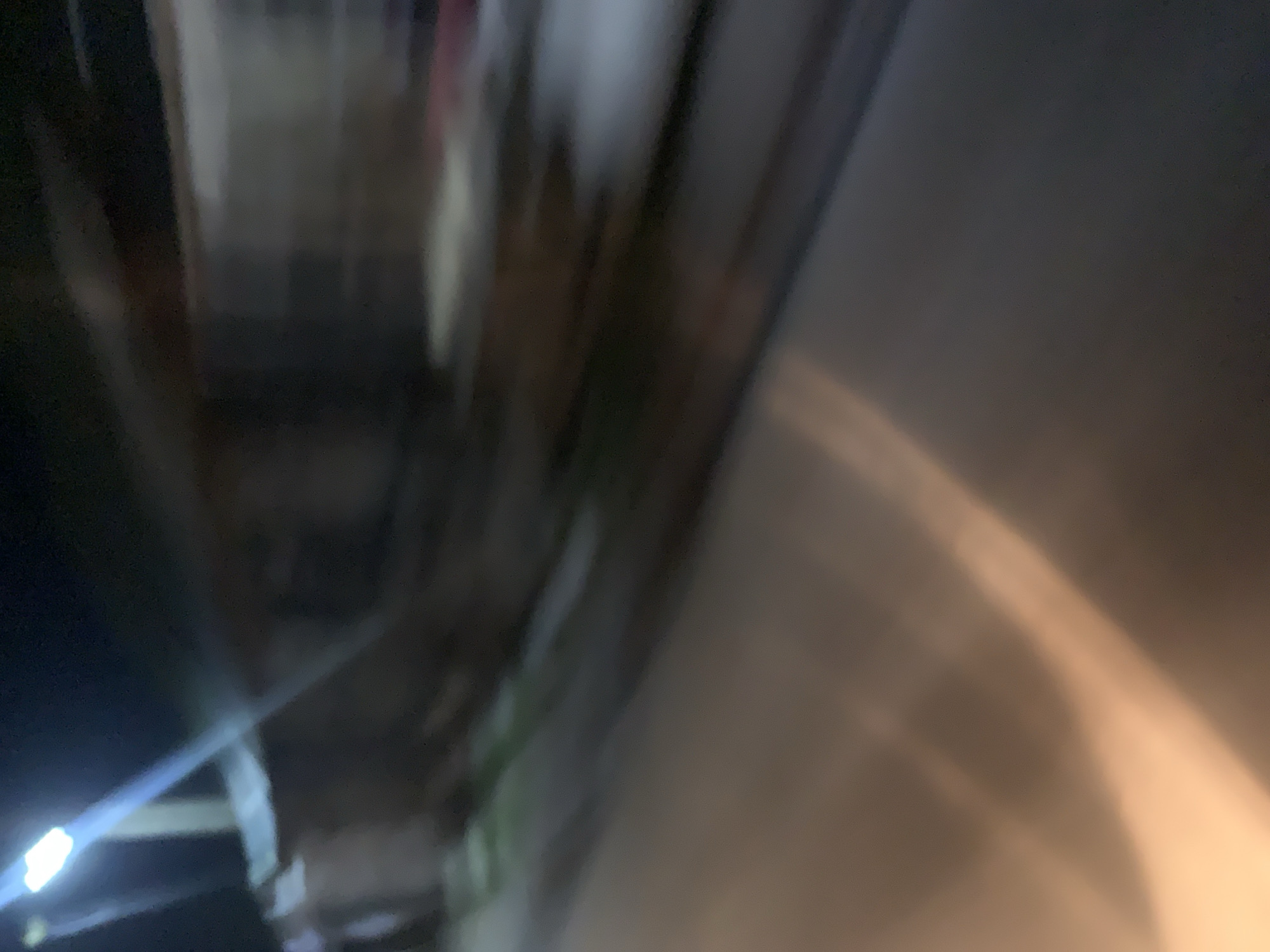 The image is a blurred, nighttime photograph taken from a moving vehicle. A bright light, possibly a streetlight or a headlamp, is visible in the upper left corner, casting a strong blue-white glare. The rest of the image is dominated by streaks of light and dark, suggesting motion blur. Hints of structures, possibly buildings or fences, are visible in the background, rendered indistinct by the blur. The foreground shows a dark, smooth surface, likely a road or pavement, with a wide, diffused band of warm light streaking across it from the left. The overall impression is one of movement through an urban or semi-urban environment at night, with limited visibility due to the motion and low light conditions.