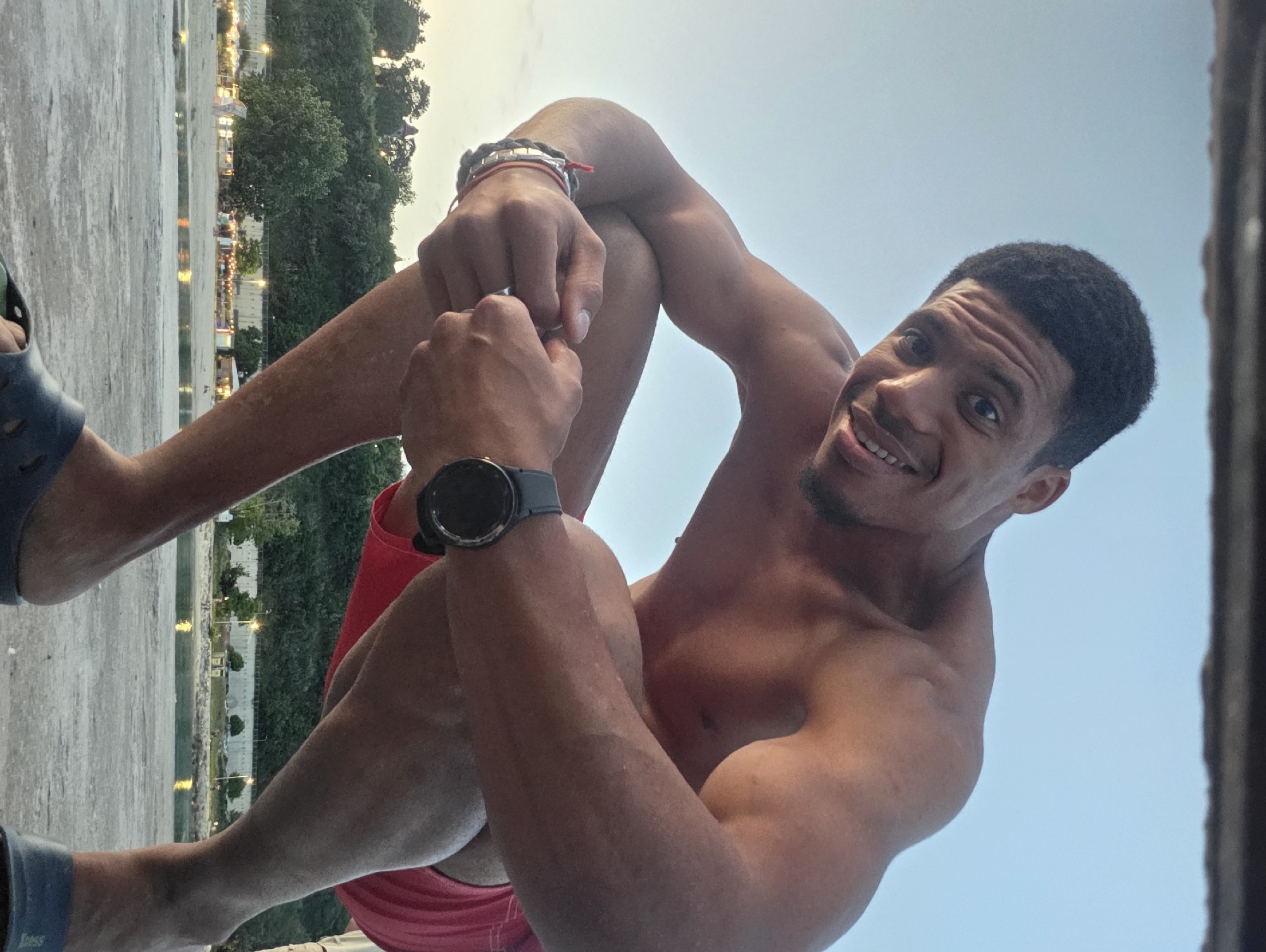 A low-angle, outdoor, waist-up shot captures a shirtless man with dark, short hair and a beard, wearing red swim trunks and a black watch on his left wrist, crouching on a concrete surface. He is looking at the camera with a wide smile, his hands clasped together in front of his knees. The background features a body of water, a sandy beach with people, buildings, and trees, under a clear sky. The overall scene is consistent with a location near the beach in Varna, Bulgaria, likely during daylight hours.