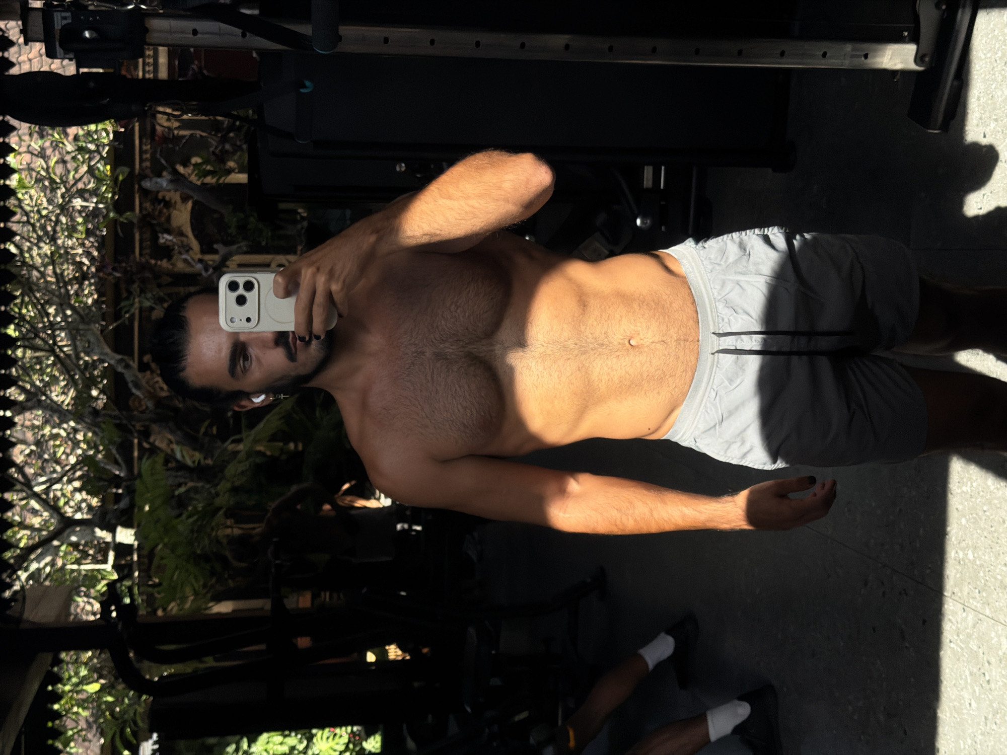 An upright, eye-level mirror selfie captures a shirtless man showcasing his physique in a gym setting in North Kuta, Indonesia. The man, with dark hair tied back and a beard, holds a light-colored smartphone with a prominent camera array in his right hand, directing his gaze towards the camera. He wears light grey athletic shorts, white socks, and dark footwear, and white wireless earbuds are visible in his ears. Notably, he has a dark manicure on his fingernails.
Strong, direct sunlight illuminates the man's left side and torso, highlighting his defined abdominal muscles and chest, and creating sharp shadows on the floor and parts of his body, suggesting it is mid-morning or early afternoon on a sunny day. The background reveals dark gym equipment, possibly racks or machinery, on the right. To the left, lush tropical foliage is visible through what appears to be a large window or open area, reinforcing the Indonesian location context. The floor is a light-colored, possibly concrete or tiled surface. Faintly visible on the elastic waistband of his shorts is the brand "NIKE."