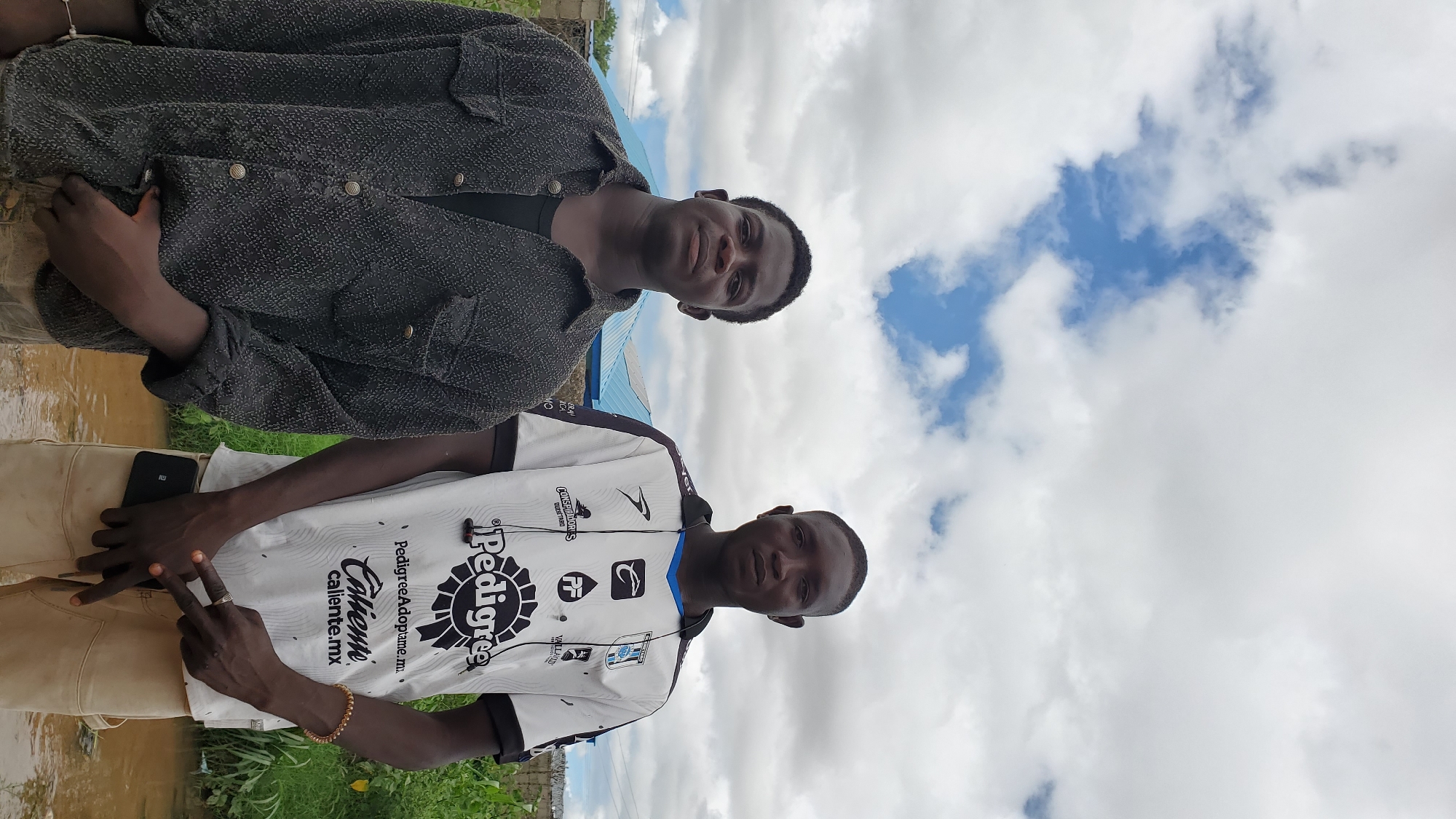 Two young Black men stand in front of a cloudy sky and a blurred background of a building and vegetation. The man on the left wears a dark, textured button-up shirt over a dark t-shirt. He has short, dark hair and is looking directly at the camera. The man on the right wears a white t-shirt with black and blue accents, featuring various logos and text, including "Pedigree" and "caliente.mx." He has short dark hair and is also looking at the camera. He is wearing light-colored pants and has a phone tucked into his waistband. Both men appear to be standing on muddy ground or shallow water. The overall scene suggests an outdoor, possibly rural or semi-urban, environment. The image was captured in Jalingo, Nigeria.