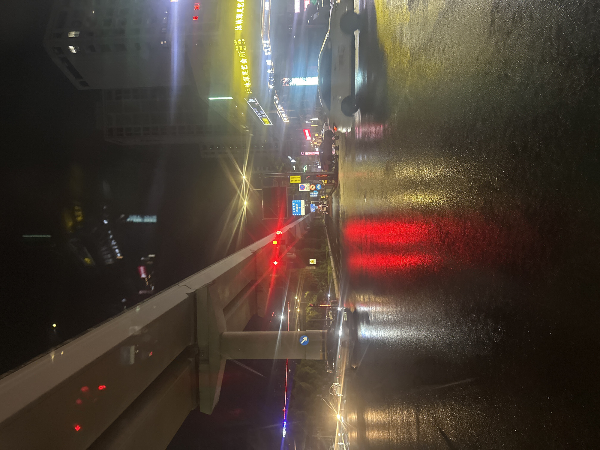 An urban night scene depicts a wet asphalt road at an intersection in the Chongqing High-tech Zone, China, under an elevated concrete highway or monorail structure. The road surface is highly reflective due to recent rain, showing vivid specular reflections of various light sources.
Dominating the left and center is a massive concrete elevated structure, supported by a thick central pillar, indicating an overpass or railway. Underneath this structure, a blue circular road sign with a white right-pointing arrow is visible.
In the mid-ground, a prominent set of red traffic lights is illuminated, casting strong red reflections onto the wet pavement. Other, less defined red lights are reflected higher up on the elevated structure itself. A yellow triangular warning sign is also present on the right side of the road.
Vehicles are in motion across the scene. A white sedan is prominently featured in the mid-right, moving from right to left across the wet intersection. Its motion blur suggests ongoing movement. A darker sedan is partially visible further back, under the elevated structure. The headlights of other vehicles are visible in the distance on the far left. A large, light-colored vehicle, possibly a van, is partially visible behind the white sedan on the far right.
On the right side of the image, multi-story buildings rise into the background. One building features a brightly illuminated yellow sign with Chinese characters, reading "珍林深足艺会所" (Zhenlin Deep Foot Art Club) and "珍林深足" (Zhenlin Deep Foot), with a smaller red "SPA" sign above it. Other building windows are sparsely lit.
Various bright streetlights and building lights cast yellow and white light, creating starburst lens flares and streaks across the image, particularly from sources on the right side. No individuals are distinctly visible, but the presence of vehicles suggests traffic activity. The overall impression is one of a bustling urban thoroughfare at night, made dramatic by the wet reflective surfaces and artificial illumination.