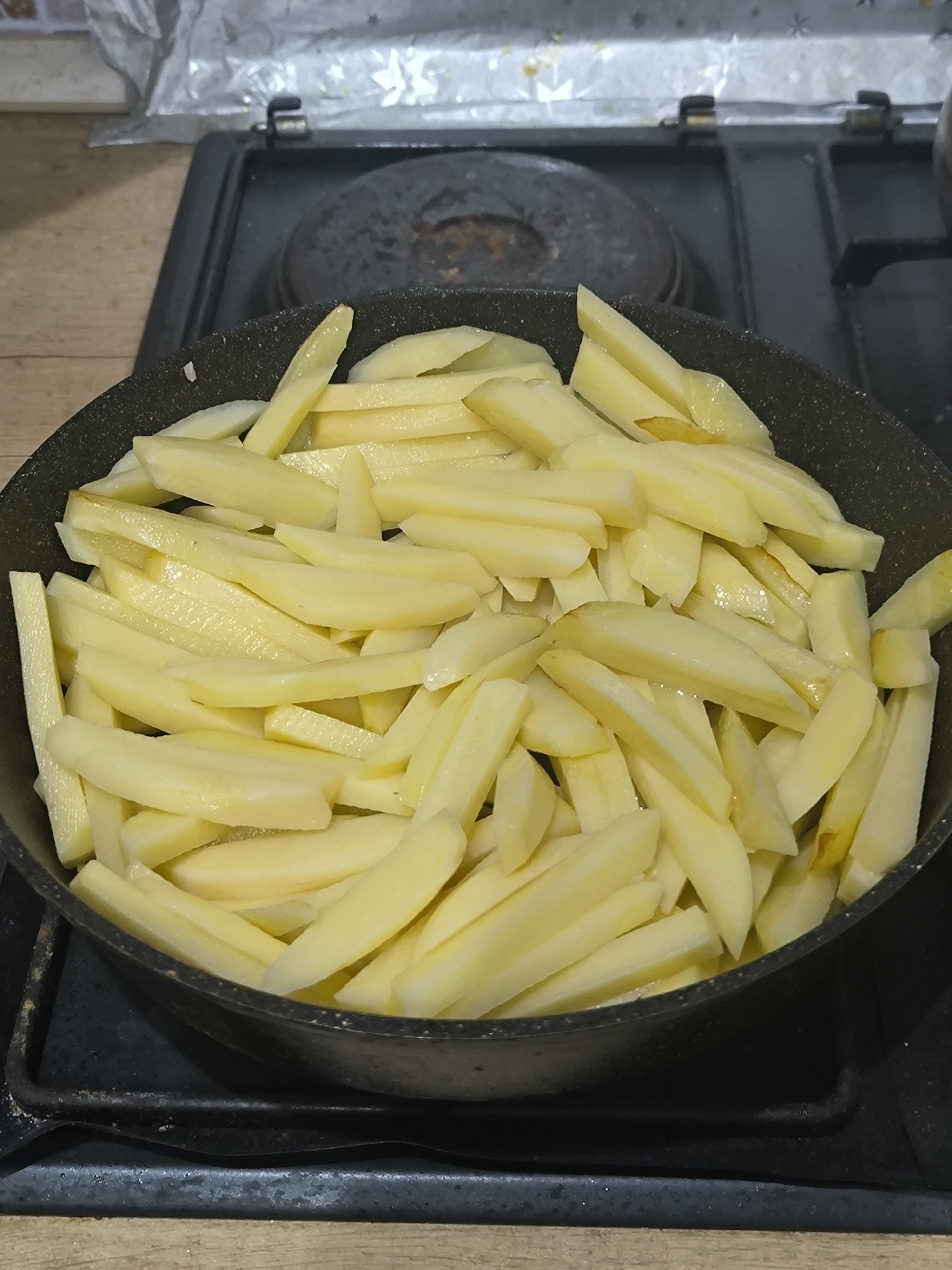 A top-down, medium shot captures uncooked, thinly sliced potatoes filling a black frying pan. The potatoes are a pale yellow and have a slightly moist sheen. The frying pan rests on a black stovetop, which appears to be a gas stove, evidenced by the burner grates. The background is out of focus, but suggests a kitchen environment with what looks like aluminum foil in the upper portion and wooden countertops or a backsplash below the stove. The lighting is slightly dim, casting subtle shadows. No people or specific landmarks are visible to suggest a location in Chișinău, Moldova.