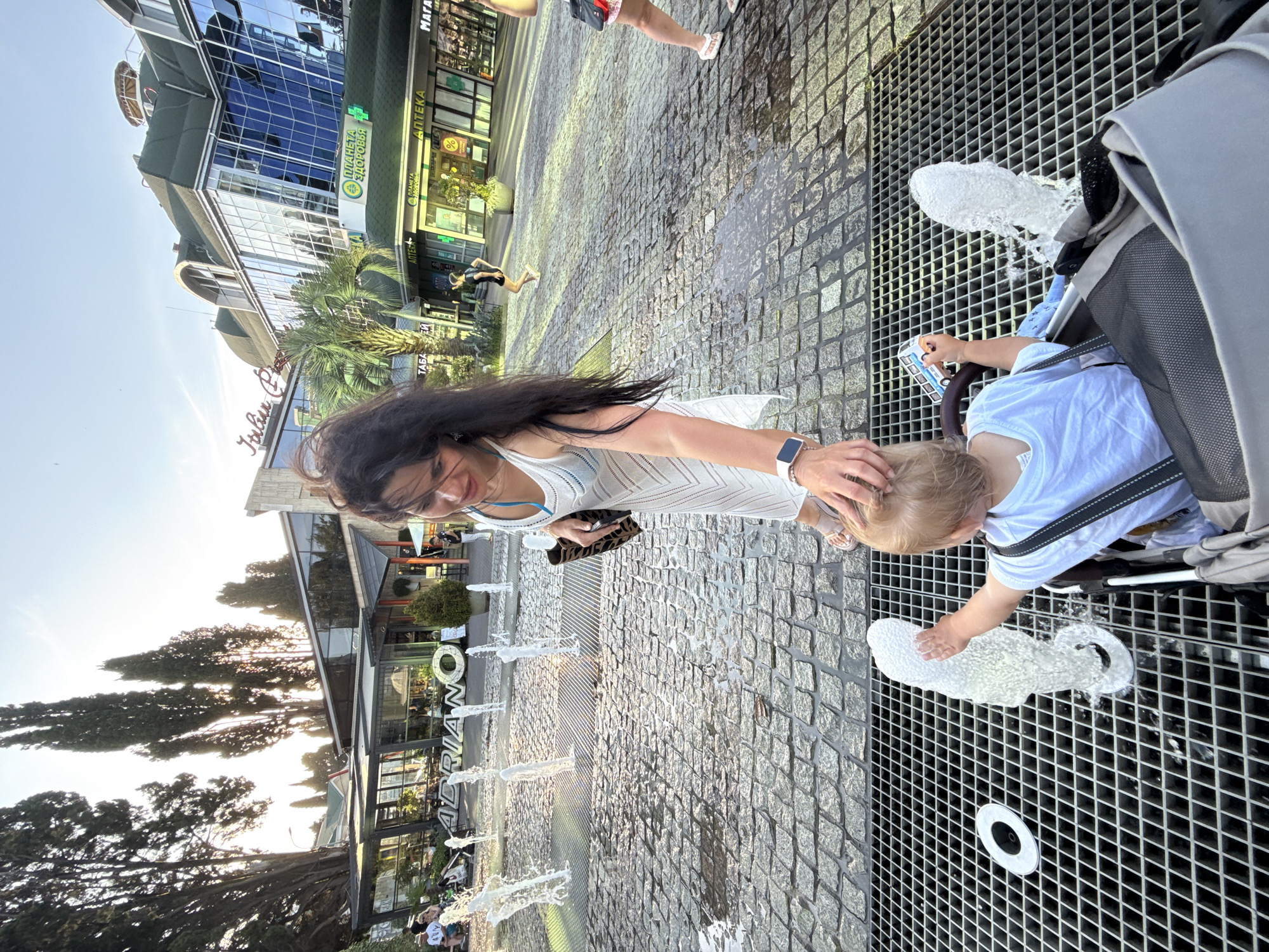In Sochi, Russia, a woman and a young child are interacting with a fountain on a sunny day. The woman, with long dark hair, is wearing a white dress and appears to be touching the child's head. The child, dressed in a white t-shirt, is seated in a stroller and is playing with the water from the fountain, reaching out a hand towards the spray. The fountain is integrated into a paved area with a metal grate. In the background, there is a modern building with large glass windows, and a palm tree is visible. Further in the distance, there are more buildings and trees, with the sun setting or rising, casting a warm light. Signs for "МАГАЗИН", "АПТЕКА", and "ПЛАНЕТА ЗДОРОВЬЯ" are visible on the buildings. Another sign with large letters spelling "ADRIANO" is also partially visible near the fountain. The overall scene suggests a leisurely outing on a pleasant day.