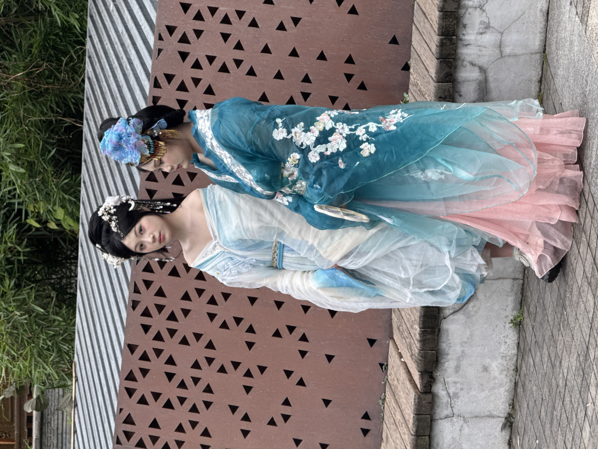 Two young women, adorned in traditional Chinese Hanfu attire, stand side-by-side in an outdoor setting. The woman on the left wears a light blue and white layered robe with flowing sleeves, her hair styled in an updo with delicate white floral ornaments. The woman on the right is dressed in a vibrant turquoise and pink ensemble, featuring intricate floral embroidery on the turquoise overlay and a voluminous pink skirt. She has a large blue floral headdress and holds a fan. They are positioned in front of a reddish-brown patterned screen, with a wooden bench and a paved walkway visible. Lush green foliage forms the backdrop. The scene appears to be in Yuzhong District, China.