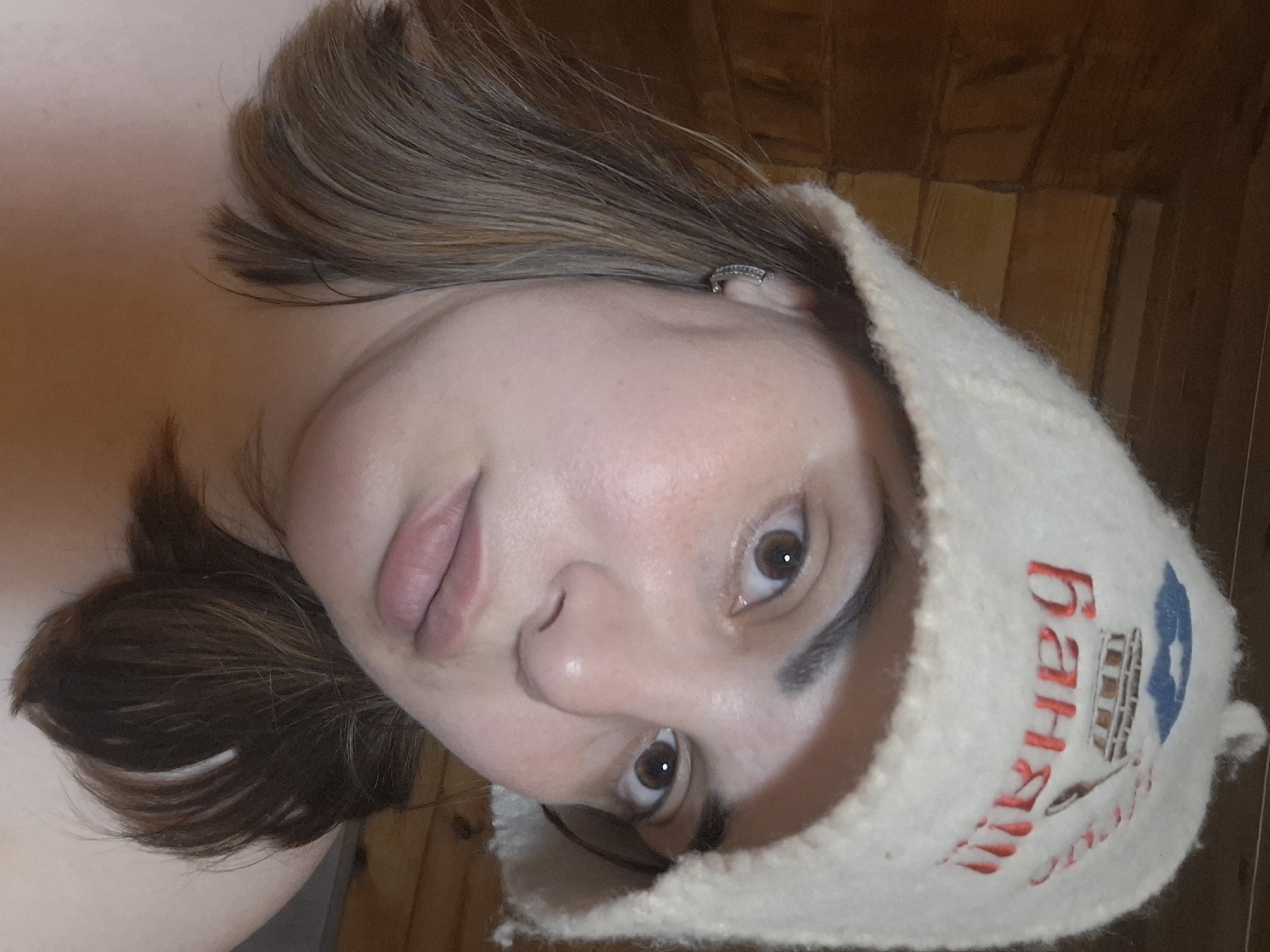 A close-up, slightly angled shot of a woman's face, who is wearing a white wool sauna hat. The hat has a design embroidered on it in red and blue. The design looks like a sauna with the word "Баня" written on it in Russian. She has brown hair and eyes. The background is blurred but seems to be wood paneling. It is plausible that the image was taken in a location in or around the city of Амзя, Russia, given the likely sauna context.