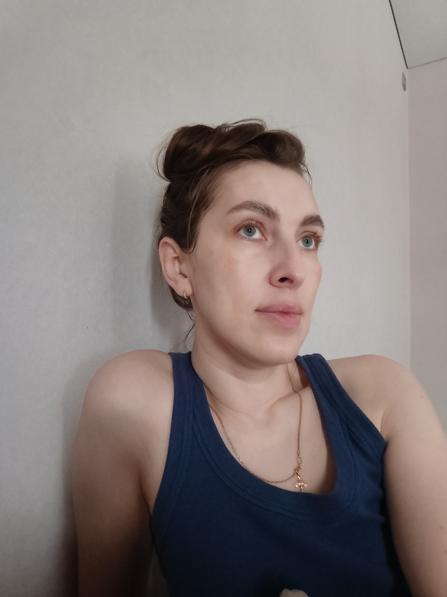 A young woman with light skin and brown hair styled in a messy bun poses in front of a white wall. She is wearing a dark blue tank top and a gold necklace with a cross pendant. The location is Chaikovsky, Russia.
