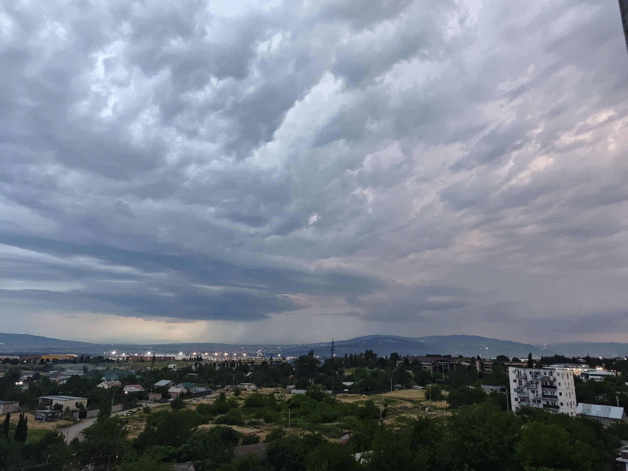 The image depicts a wide, high-angle view of a city's outskirts in Tbilisi, Georgia, under a heavily clouded sky, possibly indicating an impending storm. The foreground is dominated by lush green trees and vegetation, interspersed with a few low-lying buildings, including houses with pitched roofs and some more modern structures. A road winds through the scene. In the mid-ground, a collection of buildings, some industrial or commercial with brightly lit facades, are visible, extending towards a distant mountain range. A prominent white apartment building with multiple balconies stands out on the right side of the frame. The sky is filled with dramatic, dark grey and white clouds, with streaks of lighter sky visible on the horizon. The overall impression is of a suburban or semi-rural area on the edge of a city, with atmospheric weather conditions.