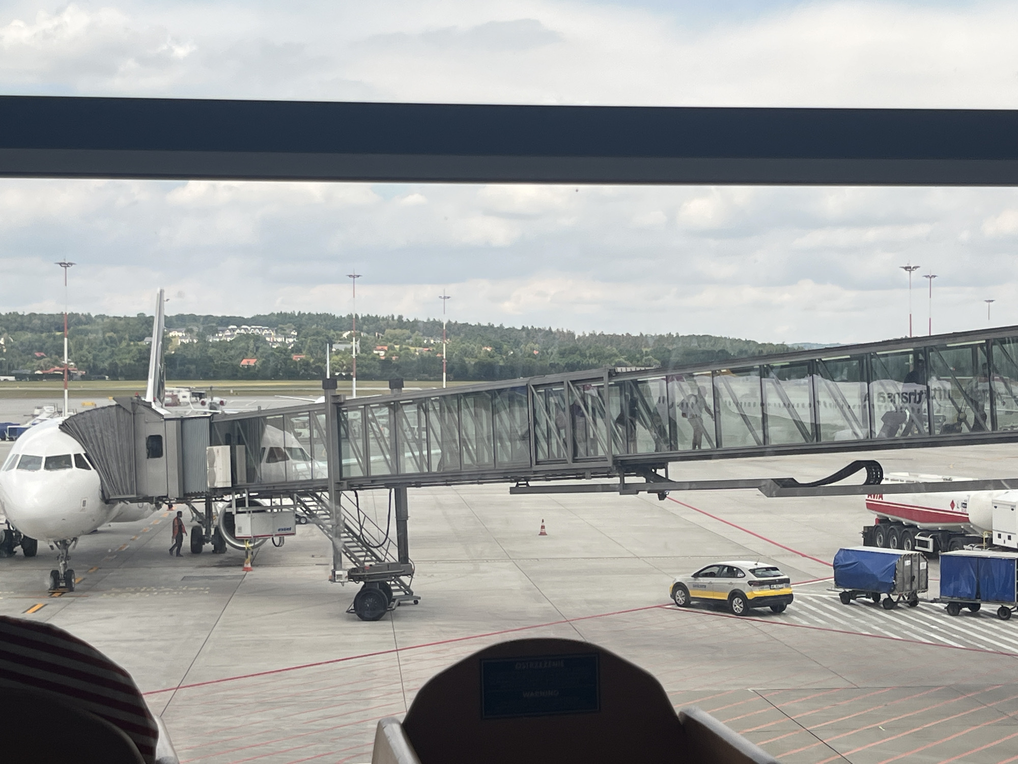 An aerial view of an airport tarmac in Balice, Poland, shows two commercial airplanes connected to a jet bridge. The image captures the scene from inside an airport terminal, looking out through a window. In the foreground, the top of two seats are visible. The tarmac area displays various airport vehicles, including a fuel truck, a small service car, and baggage carts. The sky is partly cloudy, and a green, wooded area with scattered buildings is visible in the background, suggesting the airport's proximity to a town or city.