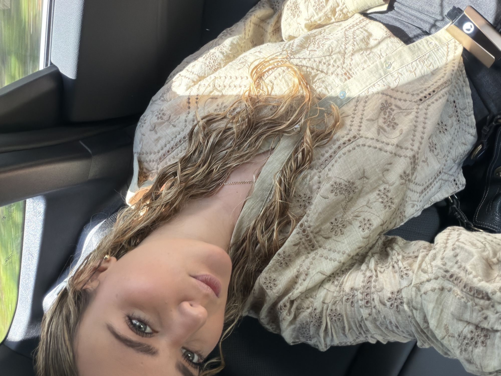 A woman with blonde, wavy hair is pictured from a tilted, close-up angle inside a car. She has fair skin, green eyes, and pink lips. She is wearing a beige, embroidered shirt with a button-down front. A thin, gold chain necklace is visible around her neck. A black purse is seen on the right side of the image, along with a metallic object. The car's interior is mostly dark gray. Outside the window, there is a blurred view of greenery, which suggests a location near vegetation. Given the prompt, this is within the city of Key Biscayne, United States of America.