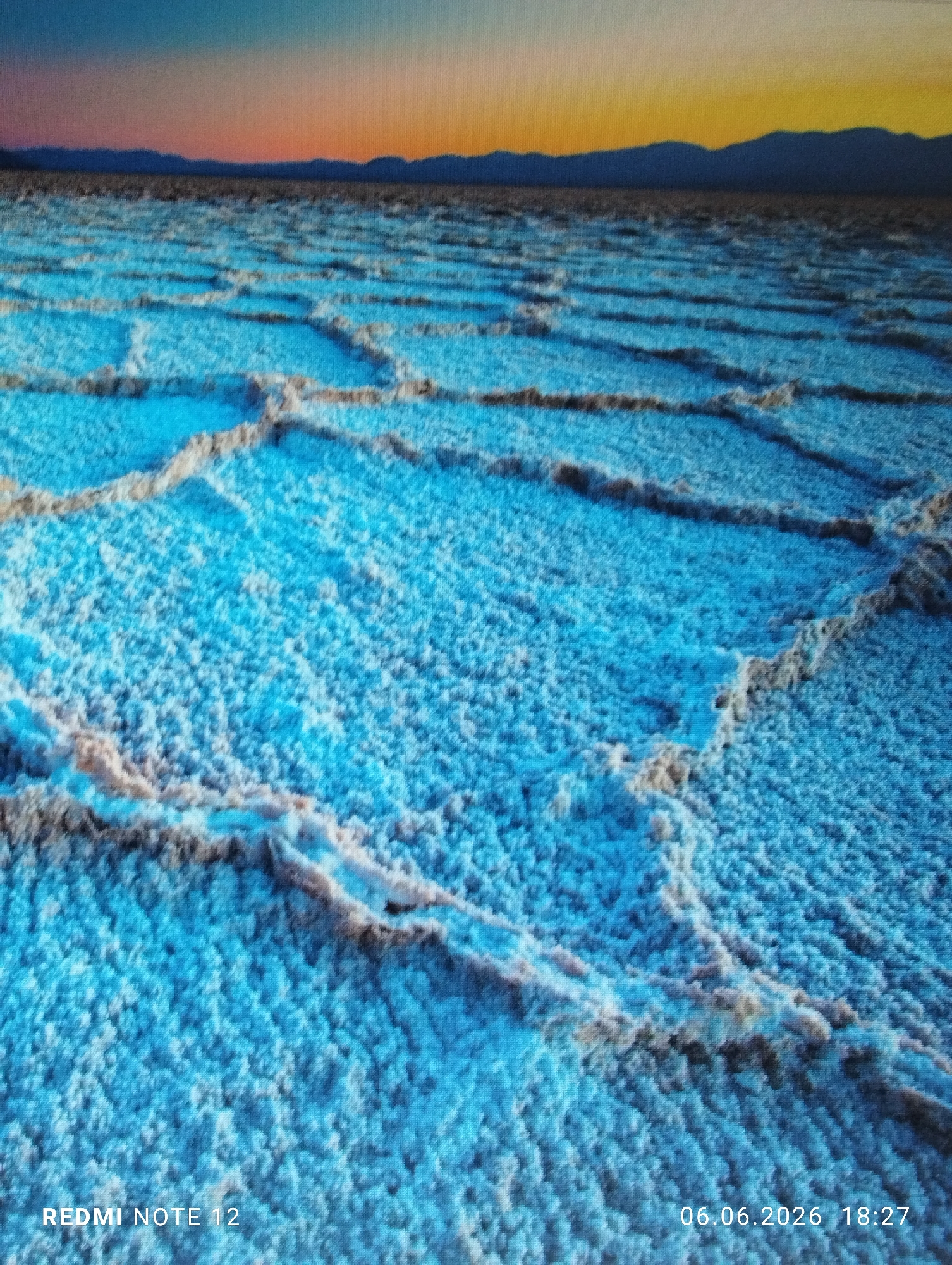 A wide-angle landscape photograph, captured in the city of Burgas, Bulgaria, depicts an expansive salt flat under a twilight sky. The foreground and midground are dominated by a textured, vibrant blue surface, characteristic of a salt pan. This blue expanse is intricately segmented by a network of raised, lighter-colored ridges, forming irregular polygonal patterns across the terrain. These ridges display a beige to off-white coloration, creating sharp contrast against the deep blue ground. The texture of the blue surface appears finely granular or crystalline. In the distant background, a dark, undulating silhouette of a mountain range or hills stretches horizontally across the frame, meeting the flat ground at a distinct horizon line. Above the mountains, the sky presents a gradient of color, transitioning from a warm orange-yellow band near the horizon, suggestive of either sunrise or sunset, to a cooler teal-blue, and then to a darker blue tone towards the top of the frame. No human figures, animals, or artificial structures are present within the depicted natural landscape. Embedded text overlays are visible in the lower corners of the image: "REDMI NOTE 12" is inscribed in white in the lower left, and "06.06.2026 18:27" is inscribed in white in the lower right.