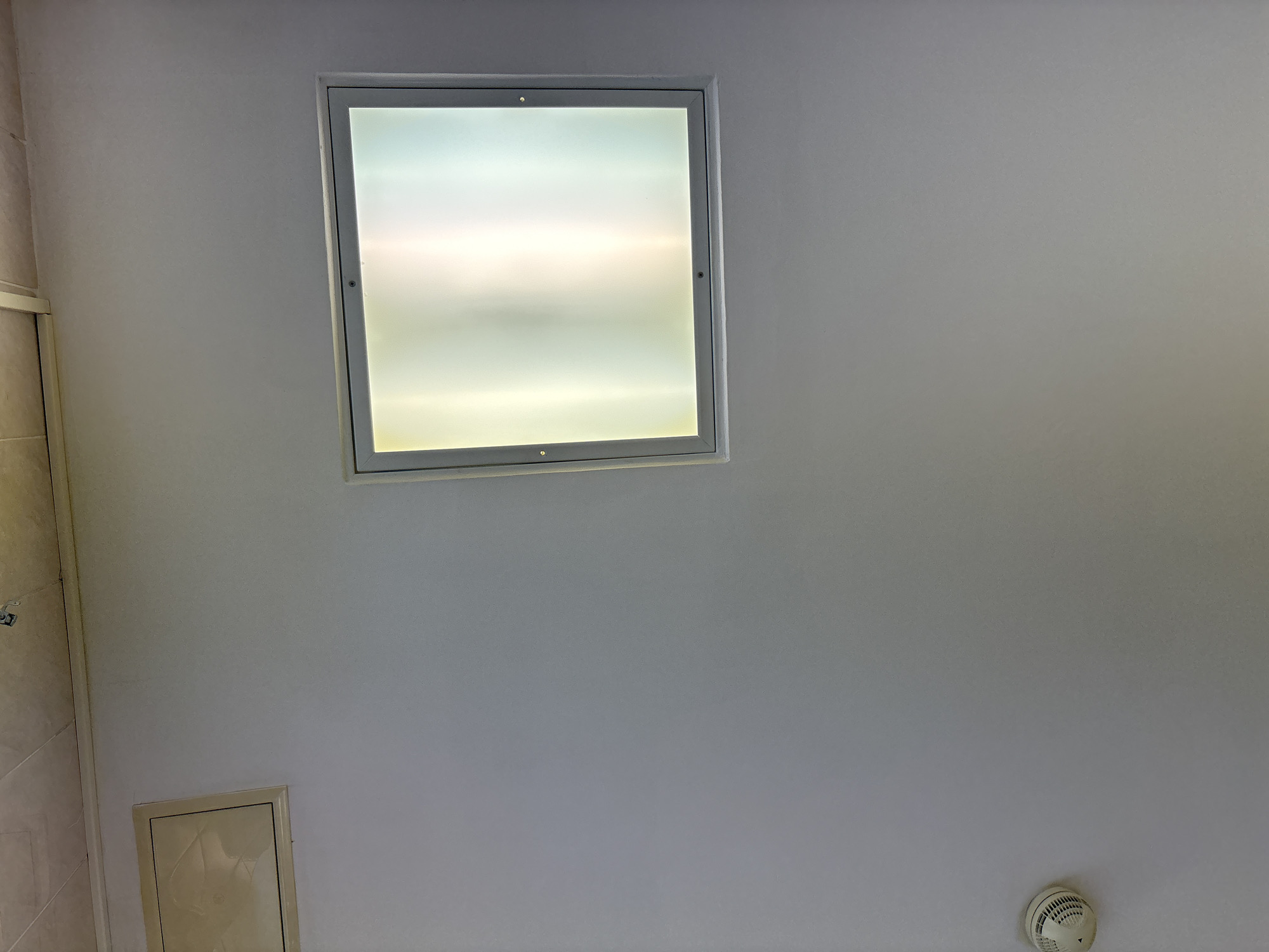 This media file captures an indoor ceiling scene, likely within a building in Saint Petersburg, Russia. The primary subject is a large, square ceiling light fixture positioned centrally in the upper half of the frame. This light features a diffused or frosted panel within a silver-toned metallic frame, emitting a bright, soft glow that shifts from a cooler white at the edges to a warmer, yellowish-white in the center.
Below and to the left of the main light, a smaller, rectangular access panel is visible on the ceiling. It is beige or light cream in color, with thin, dark lines outlining its edges and interior design details. In the bottom right corner of the image, a white smoke detector with visible grilles is mounted on the ceiling.
The ceiling itself is uniformly painted in a light, off-white or very pale gray color. A narrow strip of a tiled wall, possibly in a cream or light brown hue, is visible along the far left edge of the image. There are no people, discernible activities, or visible text in the image. The bright illumination from the ceiling light suggests it is active, providing light to the interior space. The setting appears functional and well-maintained.