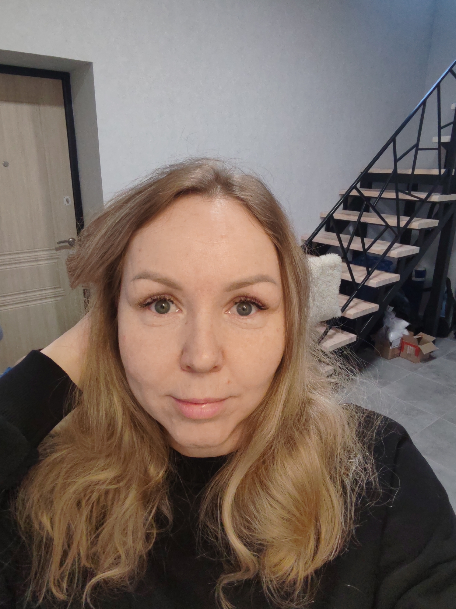 A woman with blonde hair and green eyes is shown in a close-up, indoor shot. She is wearing a black long-sleeve shirt. Her right hand is behind her head. Behind her is a door on the left and a staircase on the right with wooden steps and a black metal frame. The wall is light gray. The location is unknown. The city of Chaikovsky, Russia, cannot be confirmed from the image.