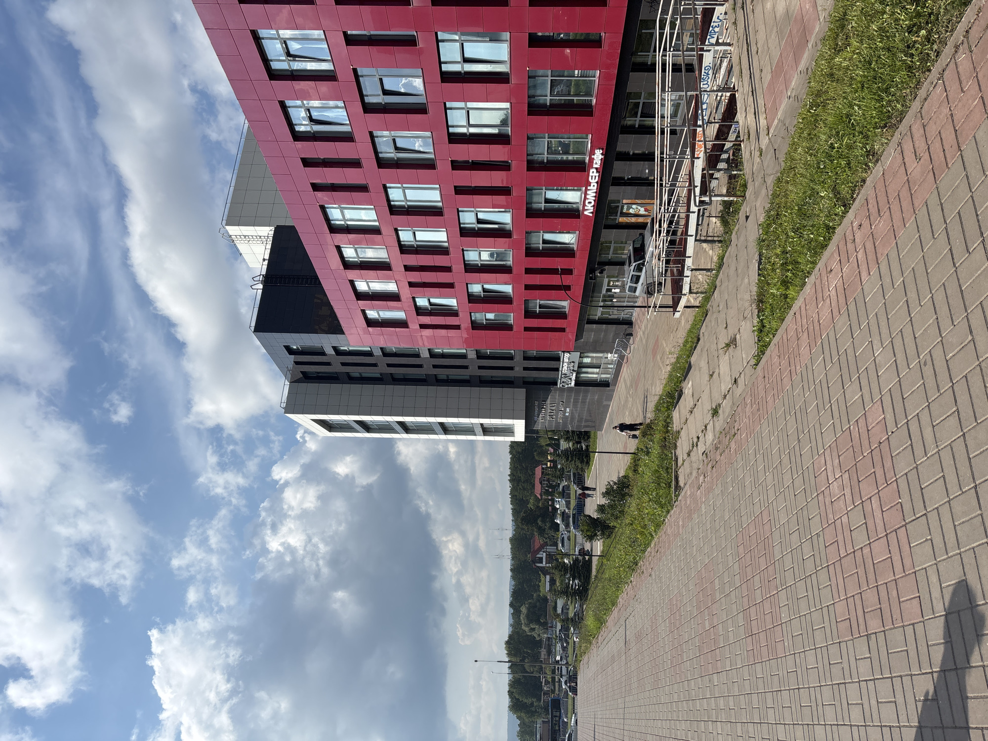 The image depicts a modern building complex on a cloudy day in Naberezhnye Chelny, Russia. The buildings feature a combination of red, black, and white facades with numerous windows. In the foreground, a red and gray brick pathway leads towards the buildings. Green grass and trees are visible along the pathway and surrounding the complex. Several cars are parked in front of the buildings, and a few people are walking on the pathway. One of the buildings has signage that reads "ЛЮМЬЕР кафе," indicating a cafe. The overall scene suggests an urban or suburban environment with contemporary architecture.