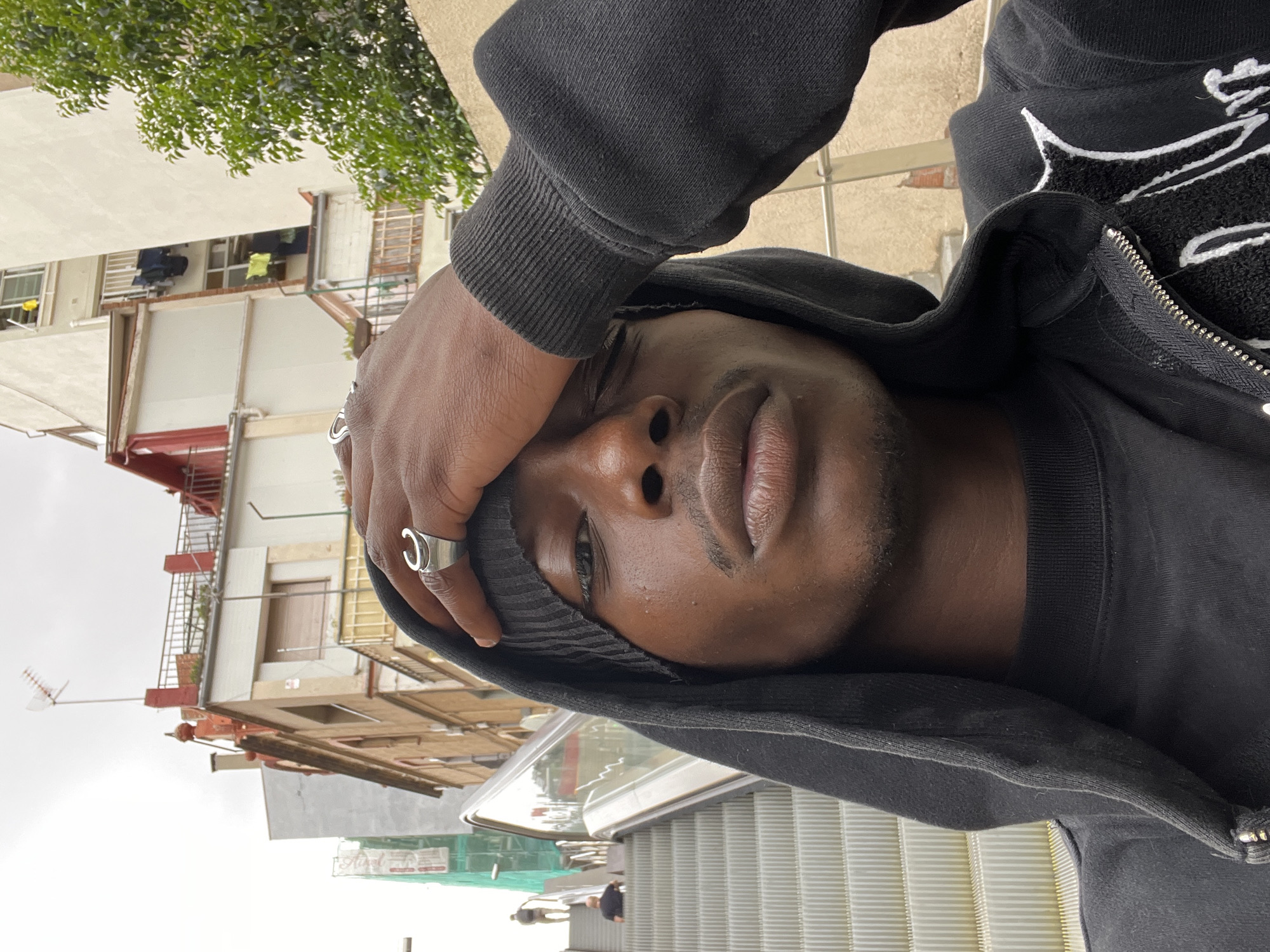 A close-up, angled, selfie-style shot features a dark-skinned male with a beanie, wearing a black hoodie, standing on an escalator. He covers one eye with his hand, which is adorned with rings. The background reveals an urban setting, specifically Barcelona, Spain, with buildings and street elements. Two figures are visible at the foot of the escalator. The weather appears overcast.