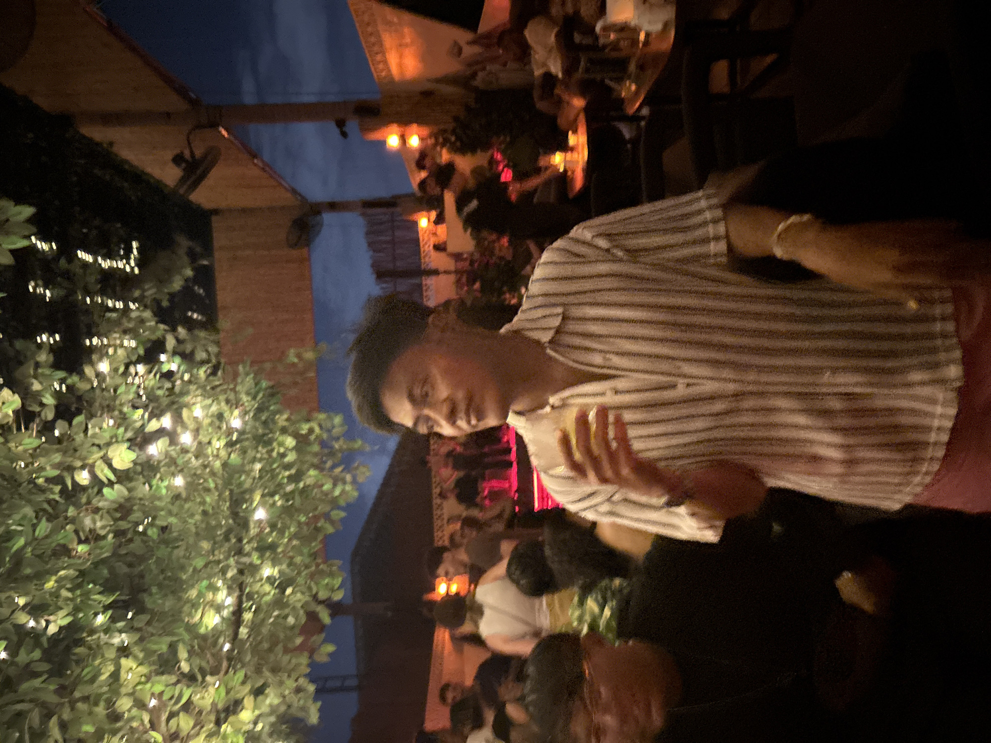 This image captures a lively evening scene at what appears to be an outdoor or semi-outdoor restaurant, bar, or lounge in North Kuta, Indonesia. The sky is dark blue, indicating it's nighttime, and the area is illuminated by various artificial lights, creating a warm and inviting atmosphere.
In the foreground, a young adult male is visible from the chest up, looking towards the left with a slight smile, suggesting contentment or enjoyment. He has short dark hair and is wearing a light-colored, short-sleeved striped shirt and possibly reddish-pink shorts. He holds a clear glass with a beverage in his left hand, and a light-colored beaded bracelet is visible on his left wrist.
The background is filled with blurred figures of other people socializing around tables, indicating a crowded gathering. Warm, orange-toned lights and string lights draped over foliage and trees contribute to the festive ambiance. To the left, a large tree or dense shrubbery is heavily adorned with numerous small, bright fairy lights, creating a sparkling effect. Overhead structures, possibly a wooden pergola or roof, are also visible, with some mounted light fixtures. The overall setting suggests a relaxed and social event, typical of a dining or entertainment venue in a tropical location. No visible text is present in the image.
