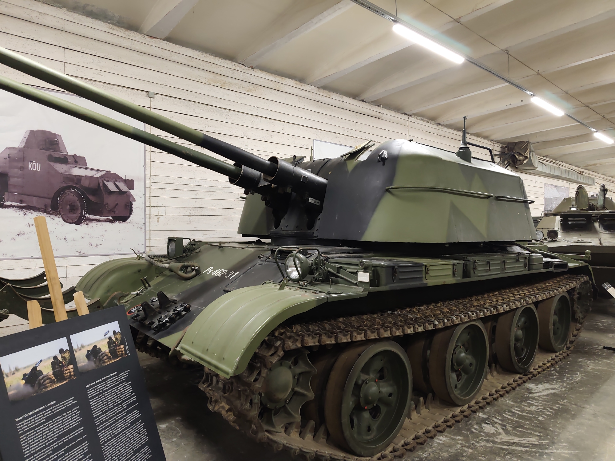 The image shows a twin-barreled anti-aircraft gun on a tracked vehicle, likely a self-propelled anti-aircraft gun, on display indoors. The vehicle is camouflaged in green and dark grey. In the background, a large photograph displays an armored vehicle with the word "KOU" visible. To the left of the anti-aircraft gun, a display board with text and images of soldiers firing a Javelin missile launcher is visible. The vehicle and display are situated in what appears to be a museum hall with a white wooden paneled wall and a concrete ceiling with fluorescent lighting. Based on the presence of an information board in Estonian mentioning "Tankitõrjesüsteem Javelin" (Anti-tank system Javelin) and the overall context of a military exhibit, this scene is likely located at the Estonian War Museum - General Laidoner Museum in Viimsi, Estonia.