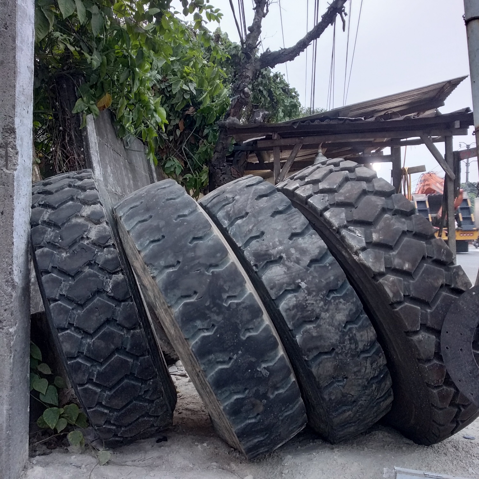 Four large industrial tires are leaning against a concrete structure in Balaraja, Indonesia. The tires are black and appear worn, with distinct tread patterns. Behind the tires, a tree with lush green leaves grows near the concrete. Further in the background, a wooden structure with a corrugated roof is visible, along with parts of an orange excavator and a yellow vehicle. The ground is covered in light-colored gravel or sand. The sky is overcast and a light gray color.