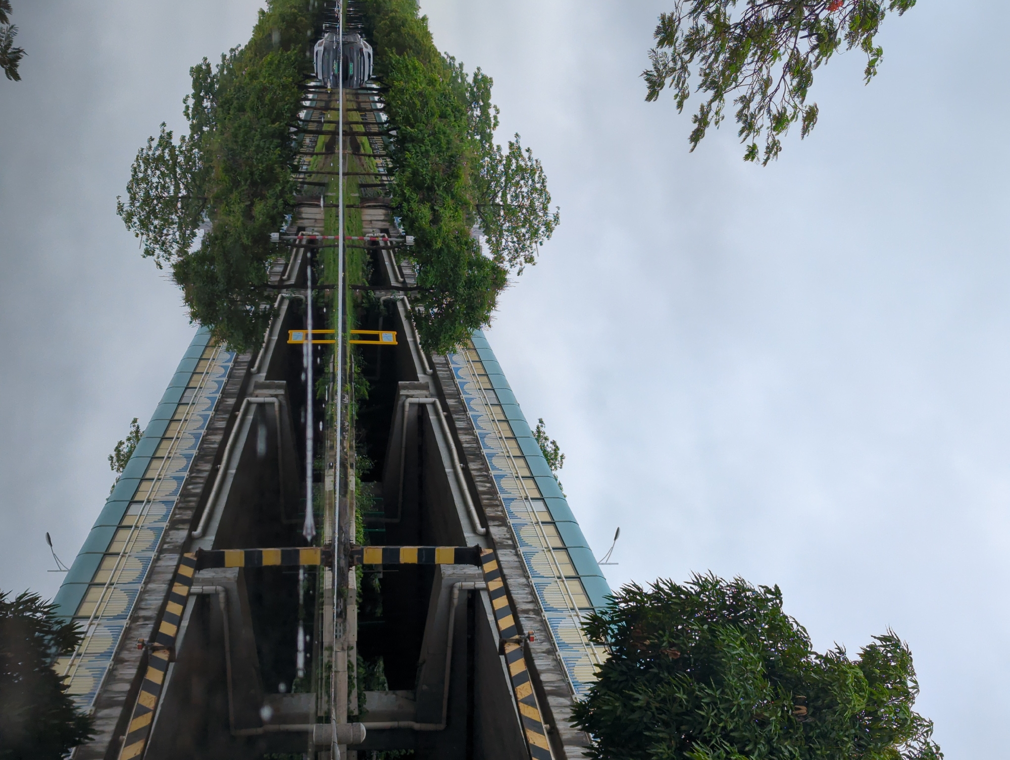 The image depicts an inverted view of an elevated structure, likely a bridge or railway, surrounded by trees. The structure has a blue and yellow facade with black and yellow striped supports. A railway track runs along the top of the structure, with a train car visible. The sky is overcast. The location is Shunde District, China.