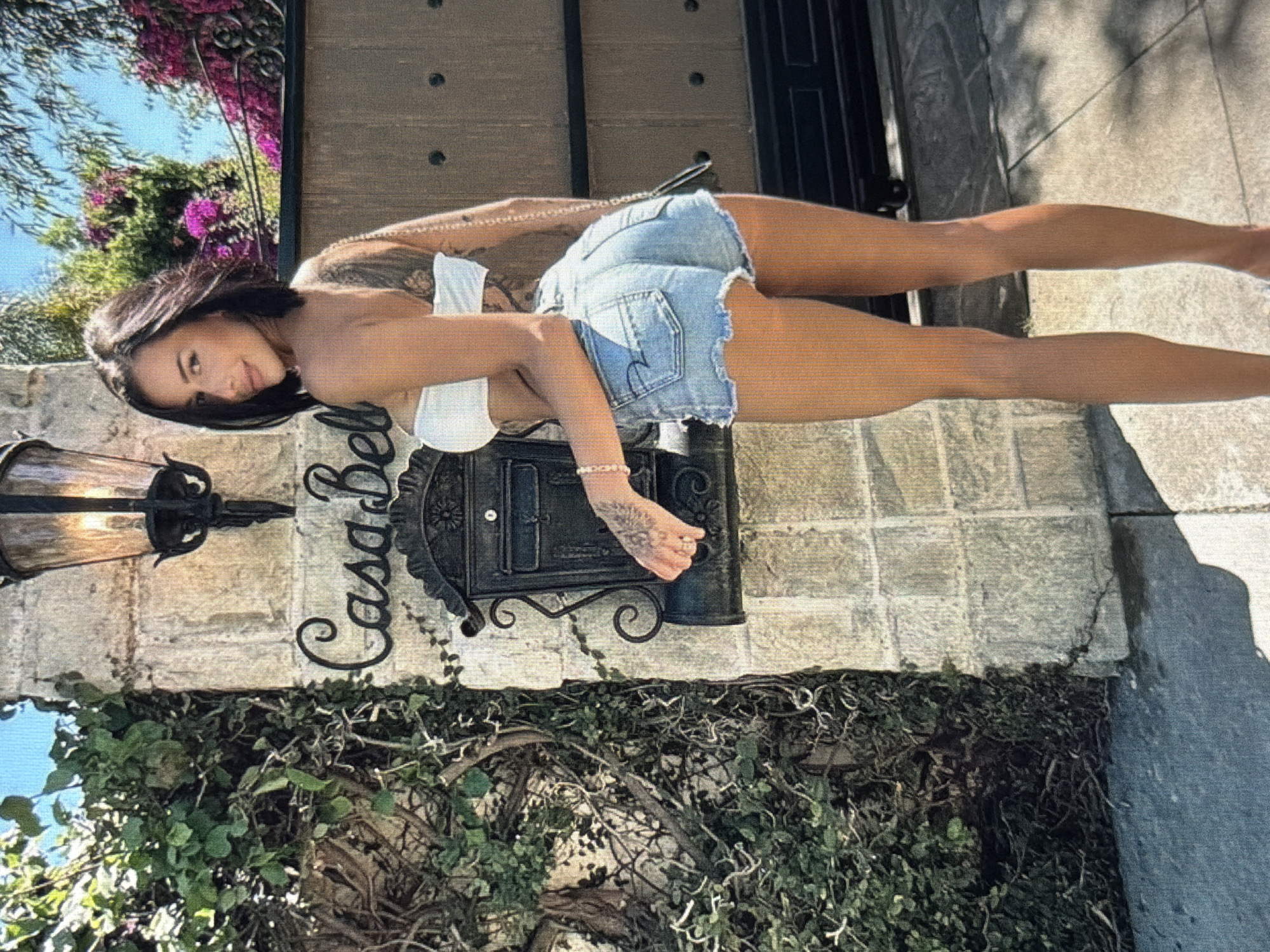 A woman with dark hair and tattoos stands outdoors in front of a stone pillar with a mailbox and a lamp. She is wearing a white bandeau top and denim shorts. Behind her is a wooden gate and lush greenery with pink flowers. The woman is facing away from the camera, looking over her left shoulder. The image is lit by sunlight. The location is not specified as being in the city of Shumen, Bulgaria.