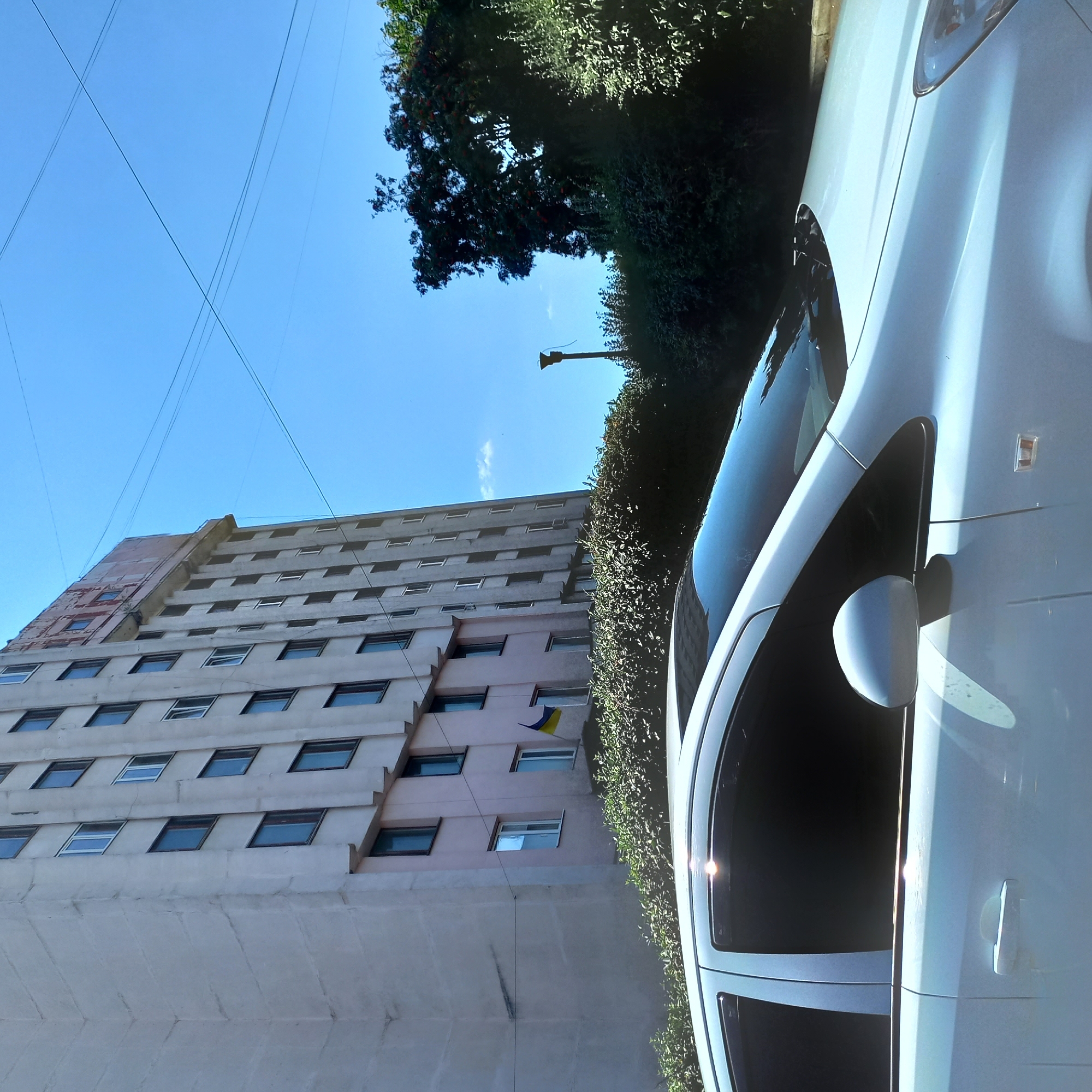 The image shows a white car parked in front of a light-colored, multi-story building. The building has numerous windows and a section under construction at the top. A Ukrainian flag is visible hanging from one of the building's windows. Lush green bushes are situated between the building and the car. Overhead, power lines crisscross the clear blue sky, and a lamppost stands to the right of the building, partially obscured by trees. The scene appears to be a typical urban street in Kropyvnytskyi, Ukraine.