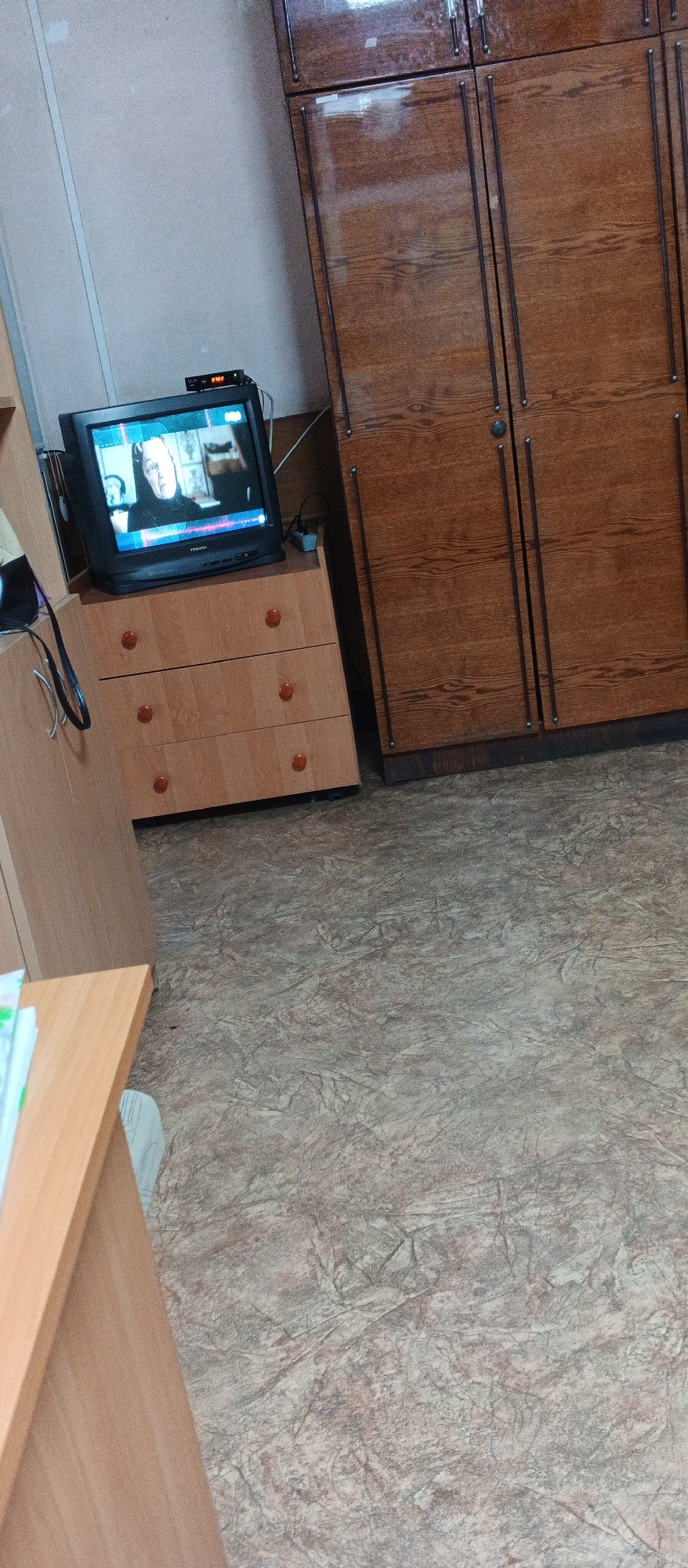 The image displays a room with a wooden dresser and a television on top of it. To the right of the dresser is a large wooden cabinet. The floor is covered in a patterned linoleum. A desk made of light-colored wood is visible in the lower left corner. The scene is likely a domestic interior or a room in an institutional setting, potentially in Smila, Ukraine.