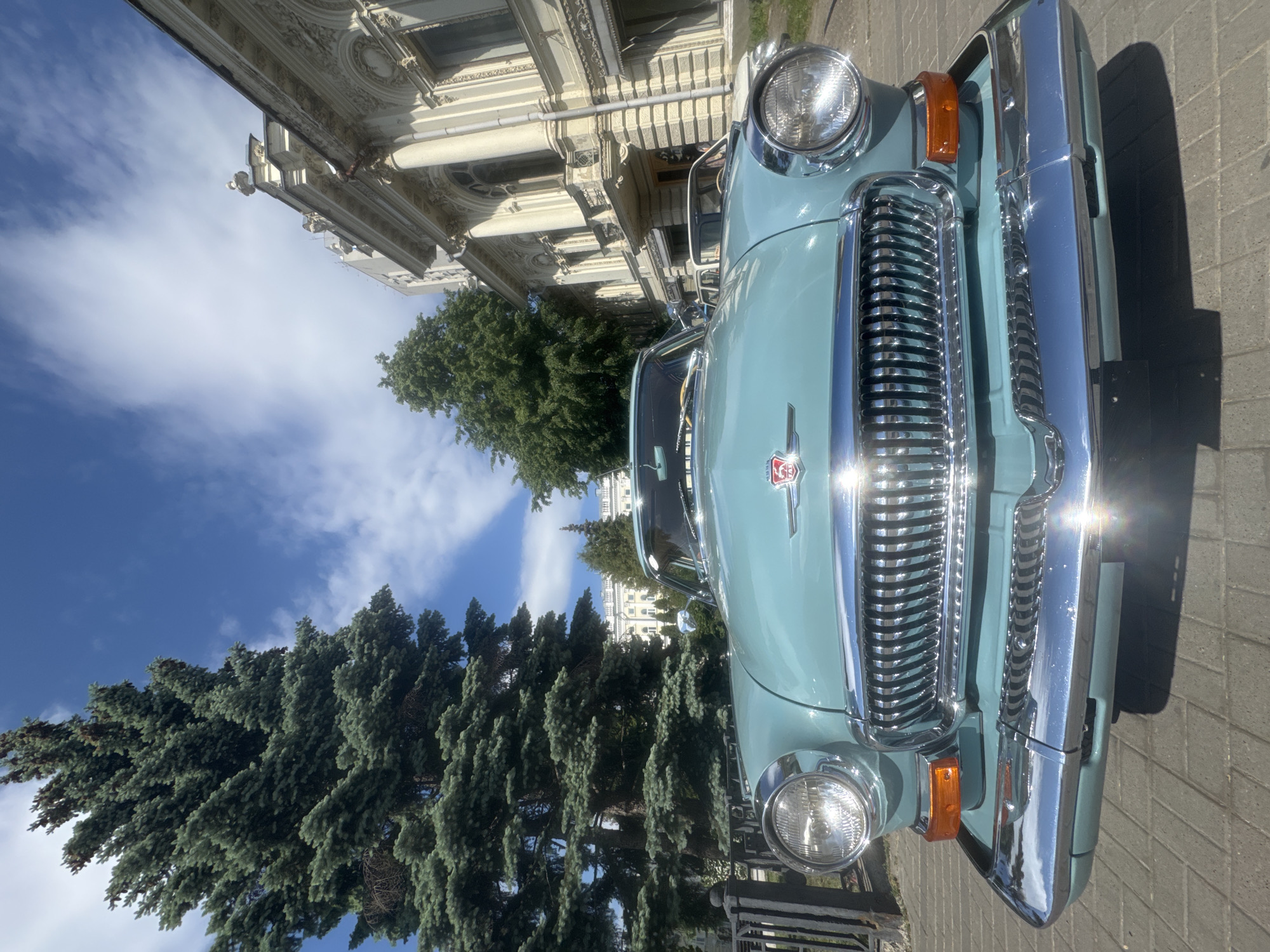 The image shows the front of a light blue vintage car with chrome grill, headlights and bumper. The car is parked on a paved surface, likely a sidewalk. Behind the car, there is a building with ornate architectural details. The building is light in color, likely white or beige. Further back, there are trees visible, and the sky is blue with some white clouds. The car appears to be a GAZ-21 Volga, and the location, given the architectural style and the car model's prevalence, is likely the city of Kazan, Russia.