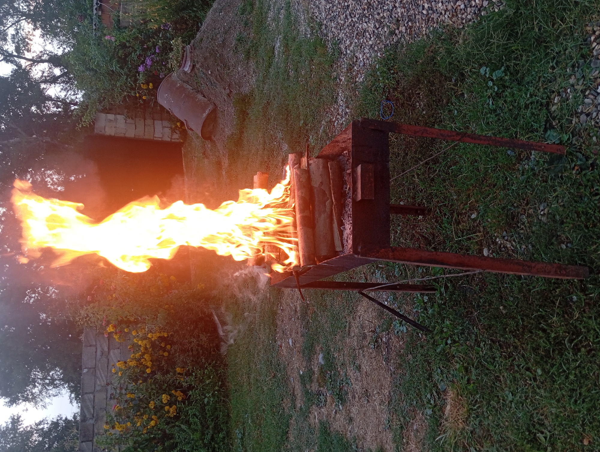 An outdoor scene depicts a lit barbecue grill with logs burning intensely. The grill is positioned on a grassy area. In the background, a stone wall borders a garden with yellow flowers. To the right of the grill, a large pile of dirt is visible, topped with a rusty metal cylinder. The overall setting appears to be a backyard or a rural area. There are no people visible in the image. The location of Şirvanlı, Azerbaijan is not evident from the visual information provided in the image.