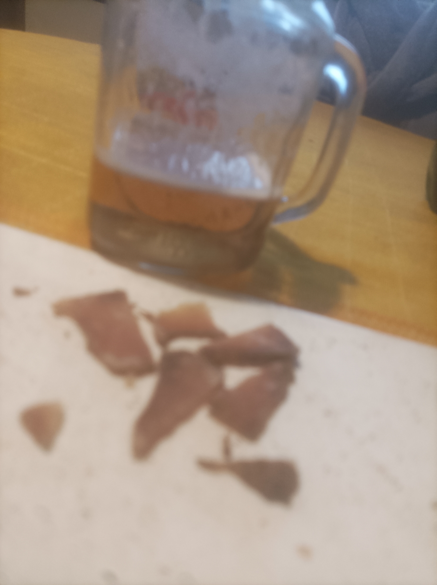 A partially filled glass beer mug rests on a light brown, possibly wooden, surface. A scattered pile of dark brown, meat-like pieces, possibly jerky, sits on a white or off-white surface in the foreground. The location and association with the city of Burgas, Bulgaria, are not apparent from the image.
