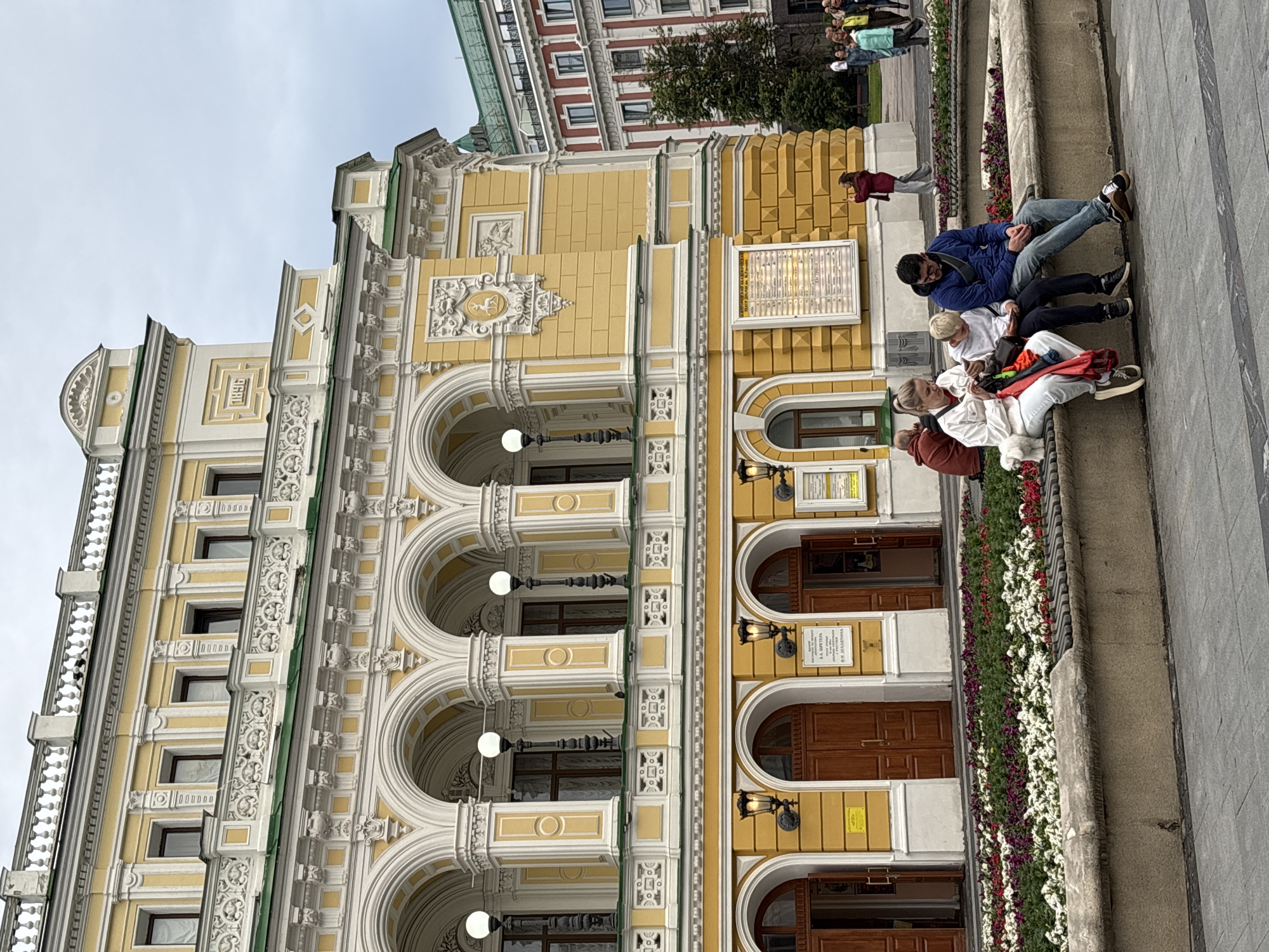 A group of people are sitting on a wide stone ledge in front of an ornate yellow building with white architectural details and arched windows. The building appears to be a grand structure, possibly a theater or historical building, with decorative elements and wooden doors. The people are casually dressed and seem to be resting or conversing. One person is wearing a blue jacket, another a white top, and a third a red garment. A fourth person is partially visible. The scene is outdoors, and the lighting suggests it could be daytime, with an overcast sky. To the right of the people, there is a flowerbed filled with colorful flowers, primarily white and purple. The ground in front of the flowerbed is paved with large gray stones. In the background, another building with red brick and many windows is visible. There is some text on plaques attached to the yellow building, but it is not legible in the image. The overall impression is a relaxed moment in a public urban space, likely a city square or promenade in Nizhny Novgorod, Russia.