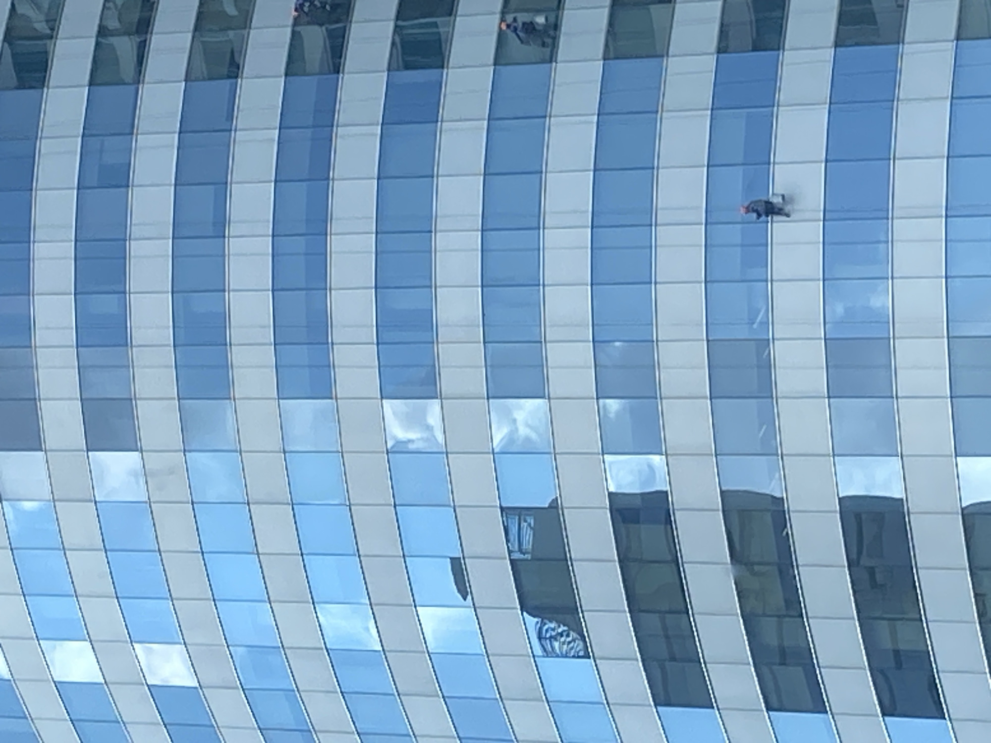 The image shows the exterior of a modern, high-rise building in Bangkok, Thailand. The building has a glass facade composed of vertical stripes of alternating white and blue-tinted windows. Some windows reflect the sky and clouds. Two window washers are visible; one near the upper part of the building and the other above the center. They appear to be suspended from ropes or harnesses, working to clean the building's exterior. The overall environment suggests a busy, urban setting.